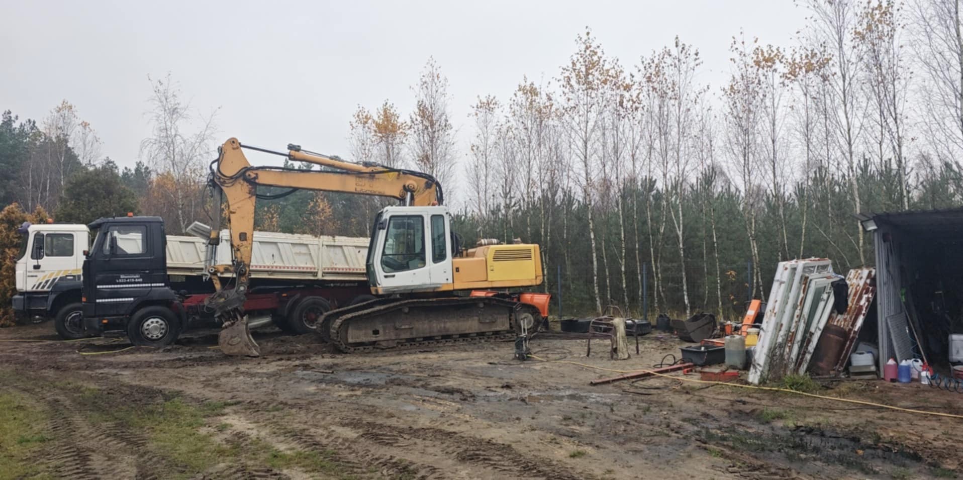 Żółto-biała koparka gąsienicowa i ciężarówka na błotnistym terenie w Śliwicach. W tle las i budynek gospodarczy z oknami. Pochmurny dzień.
