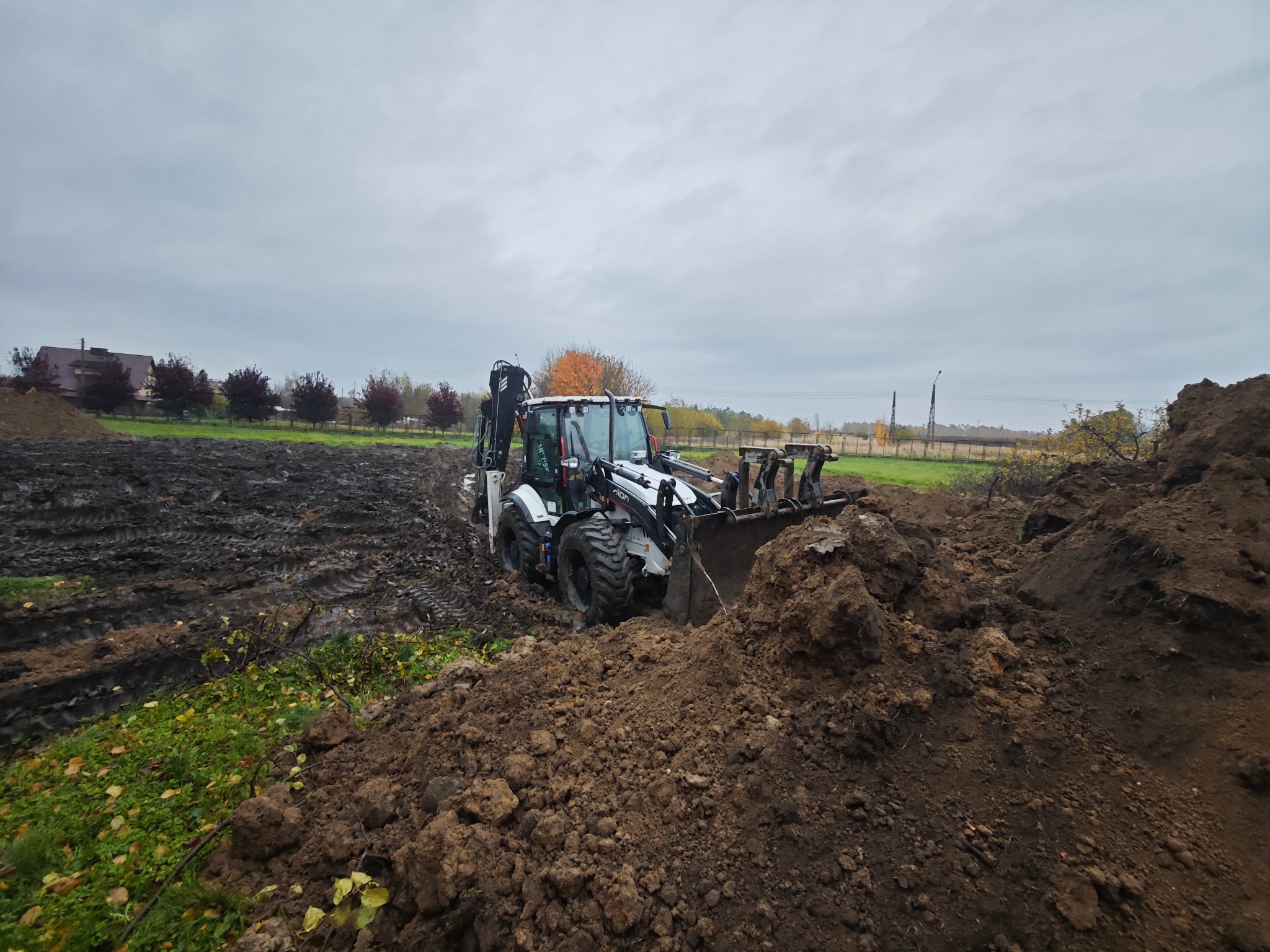 Biała koparko-ładowarka w trakcie prac ziemnych na polu, z widocznymi śladami opon w błocie i stertą ziemi. Pochmurne niebo w tle.
