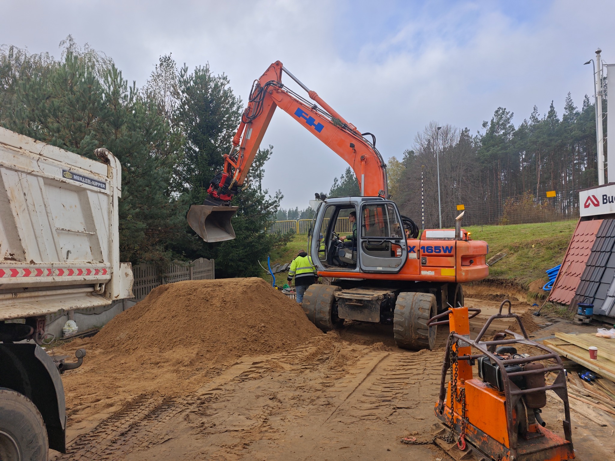 Pomarańczowa koparka kołowa FH X165W w trakcie prac ziemnych, obok wywrotka i ubijarka. W tle drzewa i budynek z ekspozycją dachówek.