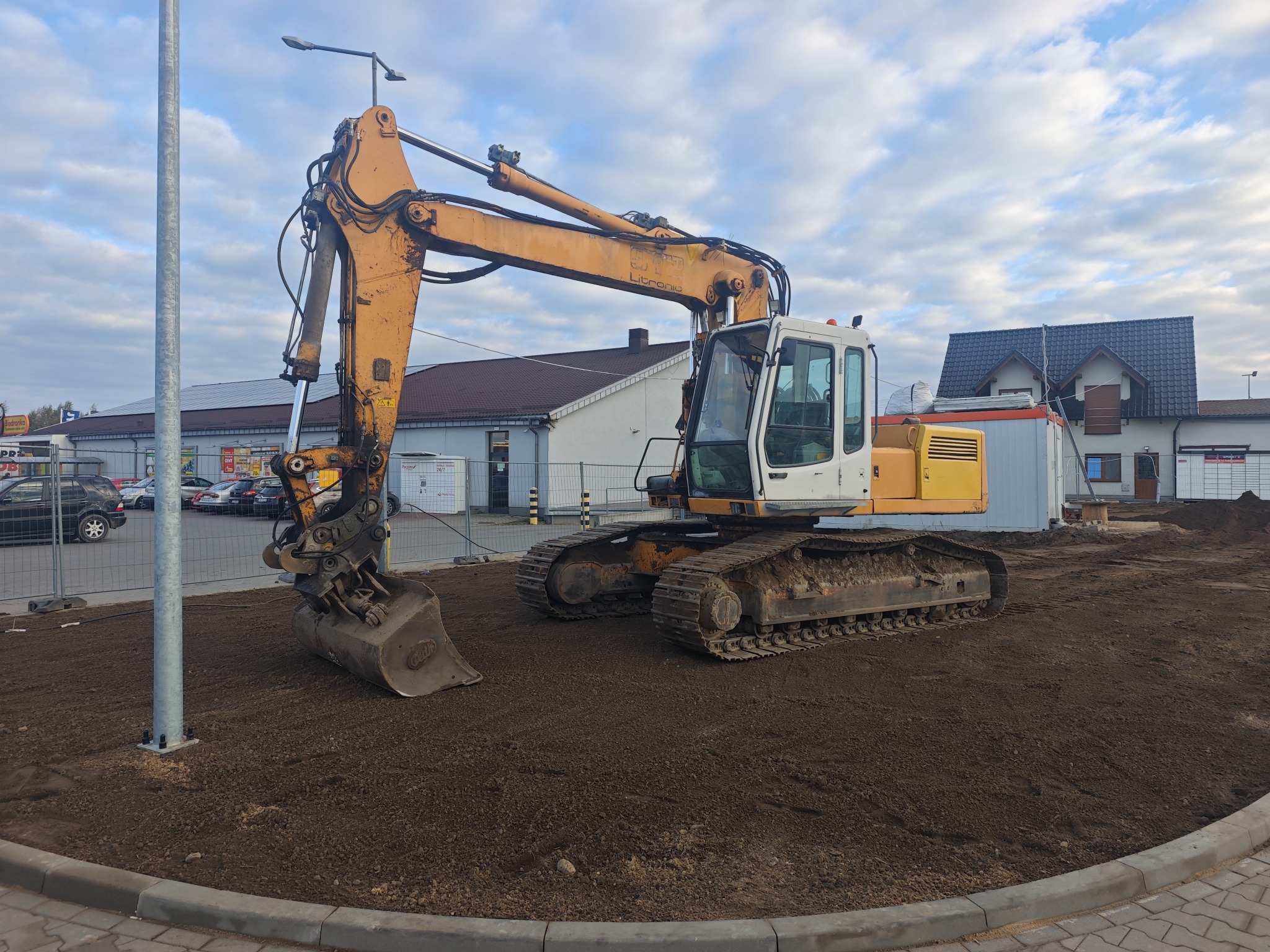 Żółta koparka gąsienicowa na wyrównanym terenie, obok słupa oświetleniowego. W tle budynki i pochmurne niebo. Prace ziemne w toku.
