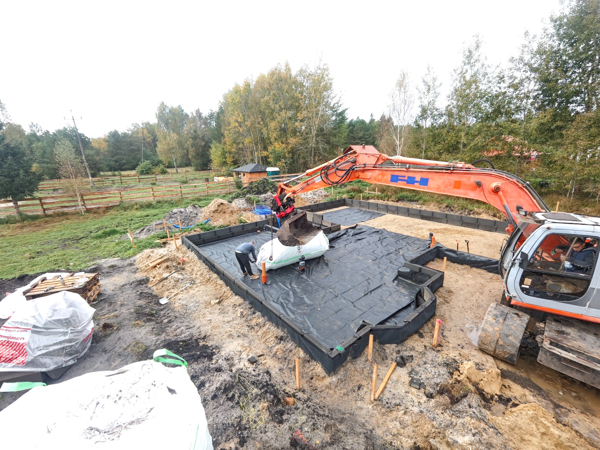 Fundamenty domu w trakcie budowy: wykop zabezpieczony czarną folią, pracownicy i koparka w akcji. Widok z góry na plac budowy w otoczeniu drzew.