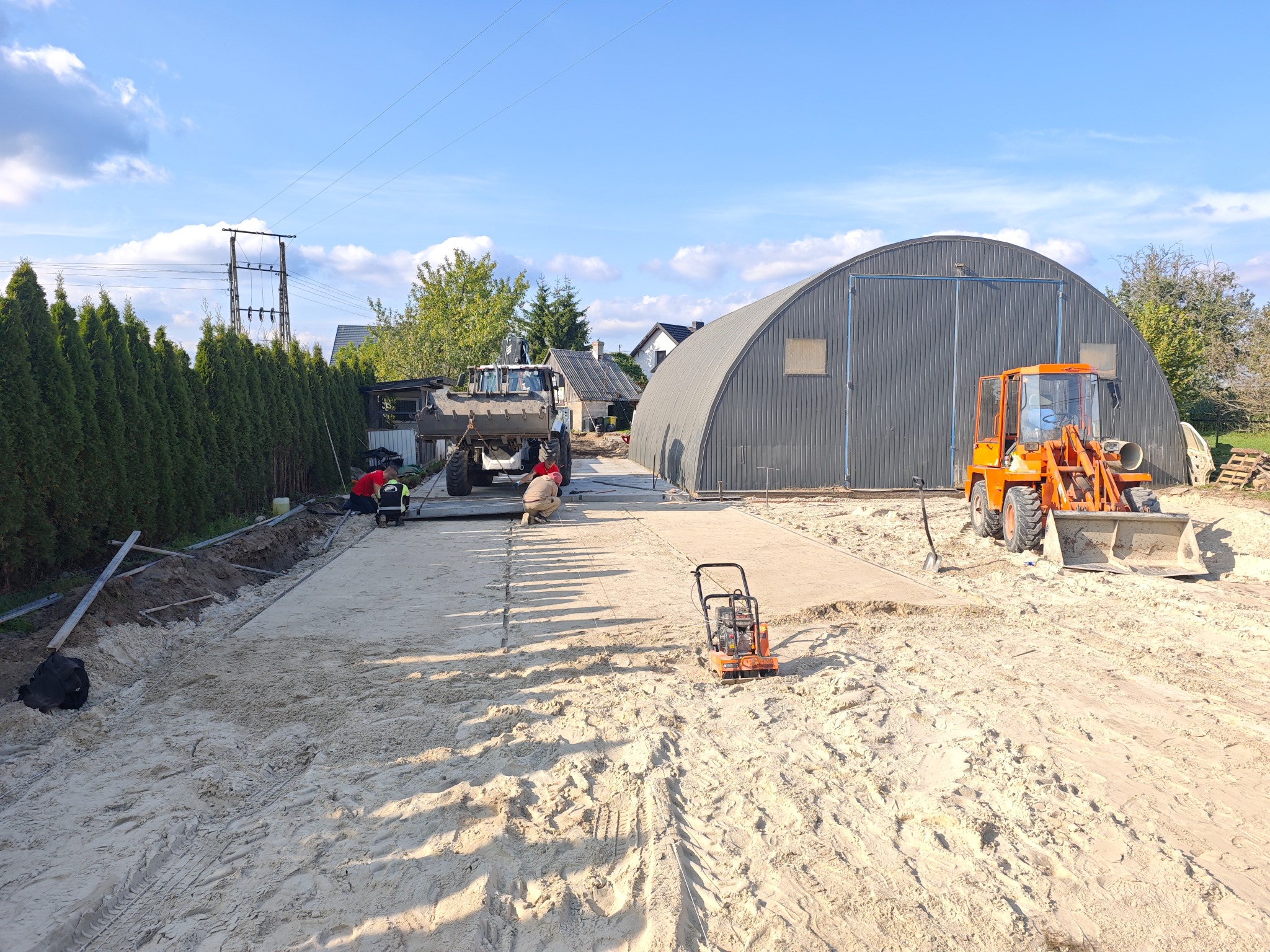 Niwelacja terenu pod budowę: ekipa wyrównuje piasek, widoczny walec, koparka i hala łukowa w tle. Prace ziemne w toku, przygotowanie podłoża pod inwestycję.