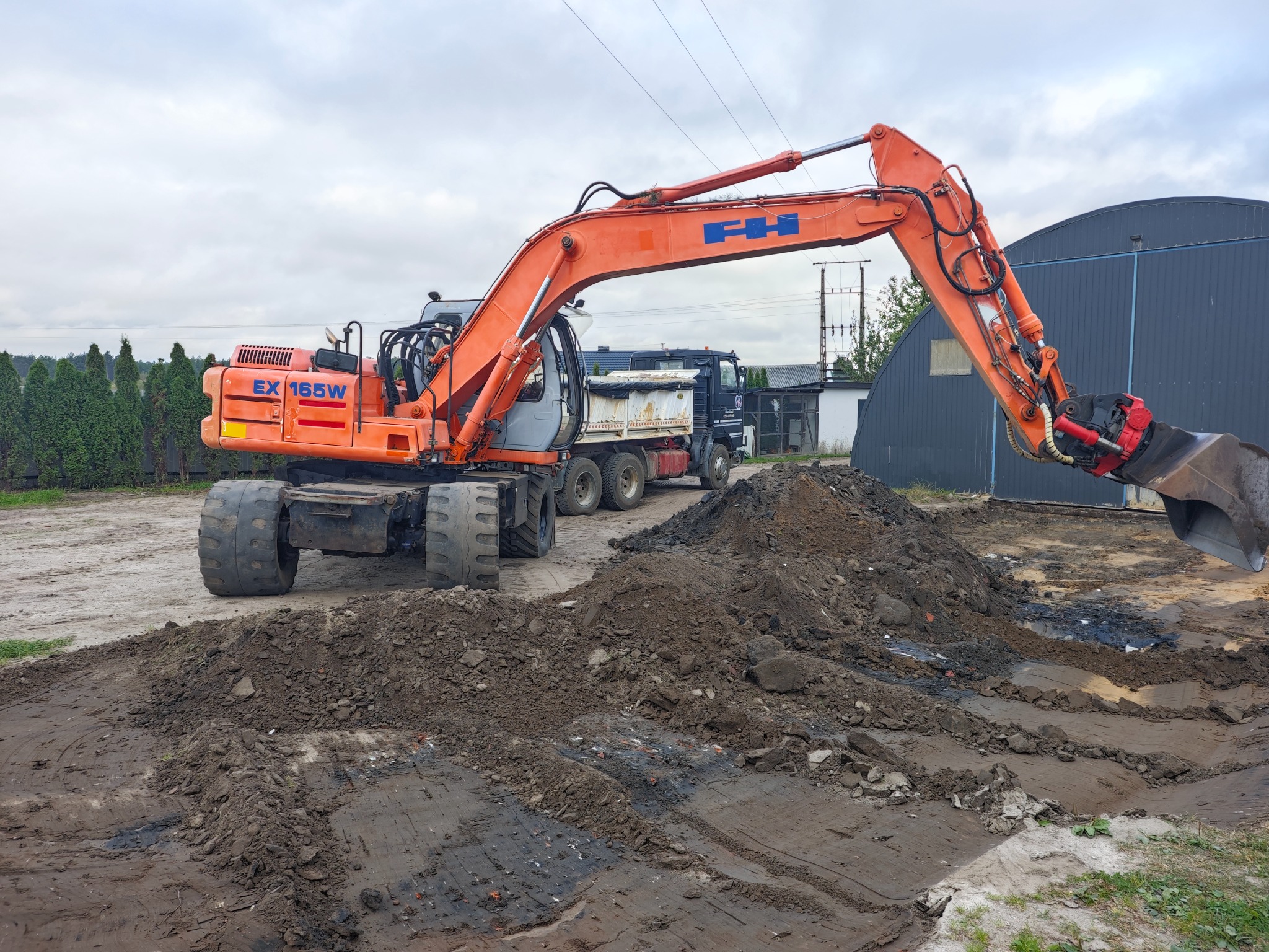 Pomarańczowa koparka kołowa FH EX 165W w trakcie prac ziemnych, z widoczną stertą ziemi i ciężarówką w tle, na tle szarego nieba i zieleni.