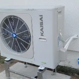 Zewnętrzna jednostka pompy ciepła Kaisai zamontowana na metalowej konstrukcji przy białej ścianie budynku, widoczne przyłącza i niebieski karbowany wąż odprowadzający skropliny.