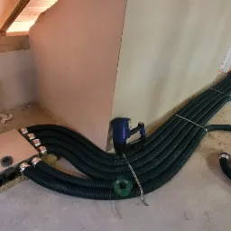 Wnętrze poddasza z widoczną instalacją rekuperacji: szereg czarnych, karbowanych rur wentylacyjnych, metalowe złącza, narzędzie do mocowania, oraz fragmenty konstrukcji drewnianej i ocieplenia.