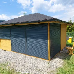 Drewniana altana z zasuwanymi roletami w kolorze grafitowym, pokryta czarnym gontem bitumicznym, otoczona zielenią i górskim krajobrazem w tle.