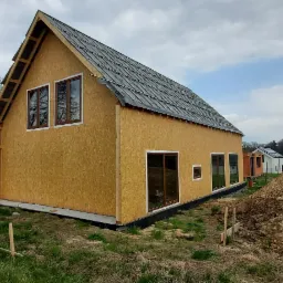 Dom w trakcie budowy z drewnianą konstrukcją szkieletową, widoczne okna z brązowymi ramami i częściowo pokryty dach, na pierwszym planie hałda ziemi.