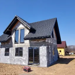 Surowy stan domu jednorodzinnego z szarą dachówką, widoczne okna i drzwi w ciemnych ramach, teren wokół domu w stanie przygotowania pod ogród, w tle drugi budynek i drzewa.