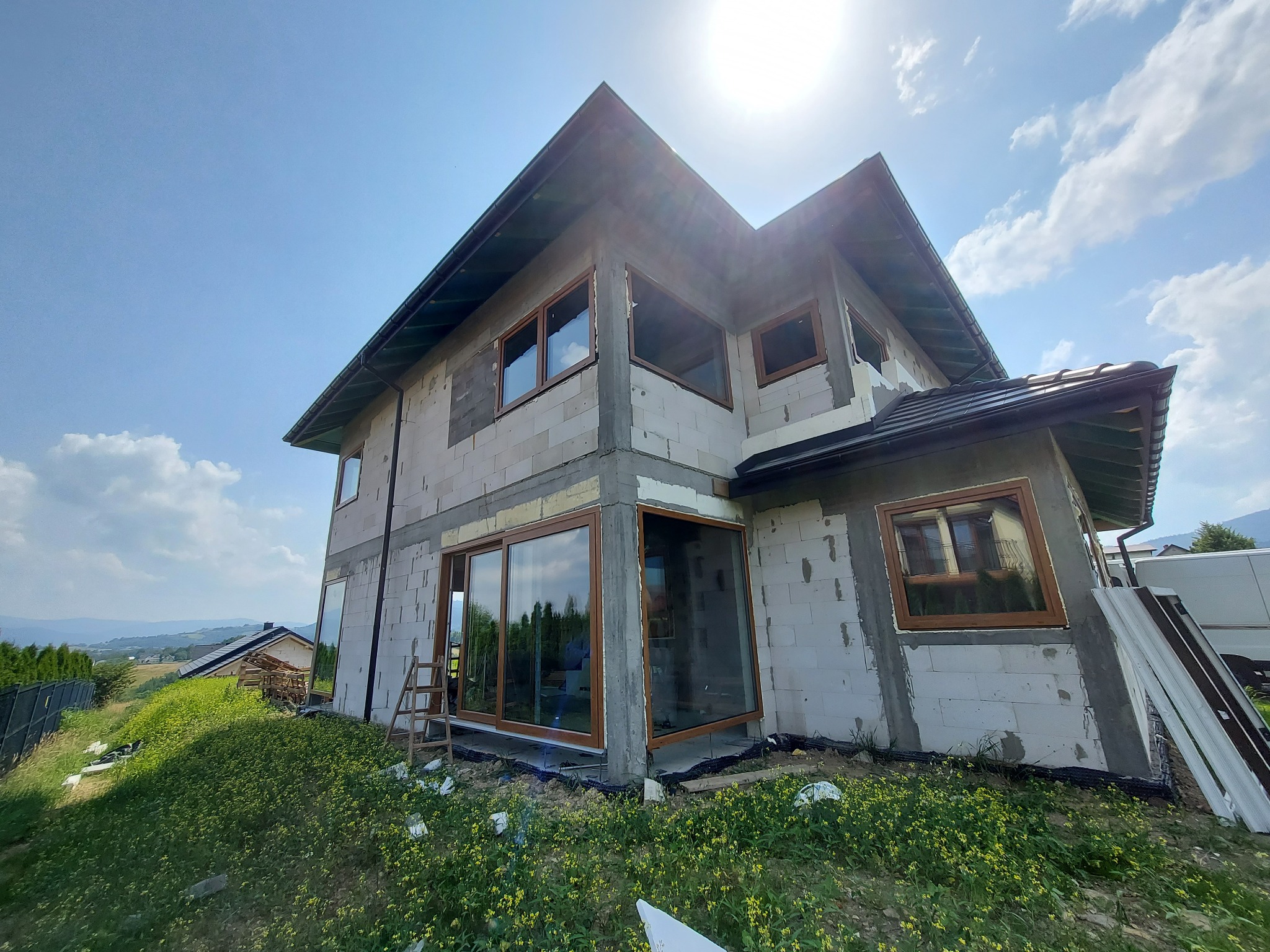 Surowy stan domu jednorodzinnego z widoczną konstrukcją ścian z bloczków, częściowo zamontowanymi oknami z brązowymi ramami i rozsuwanymi drzwiami tarasowymi, drabina oparta o ścianę, widok...