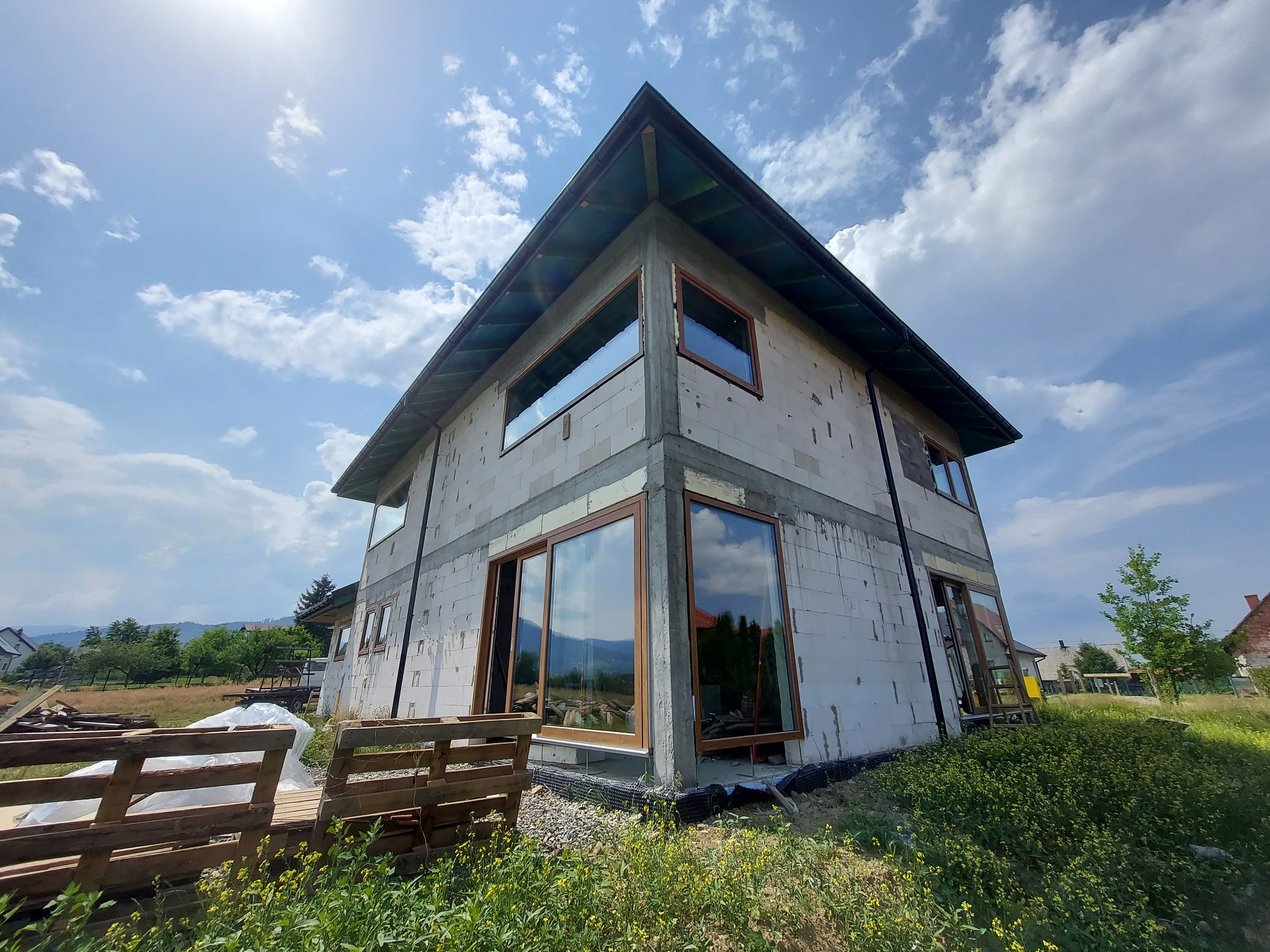 Surowy stan dwukondygnacyjnego domu z zamontowanymi oknami i drzwiami przesuwnymi, widok z dołu na tle błękitnego nieba z chmurami.