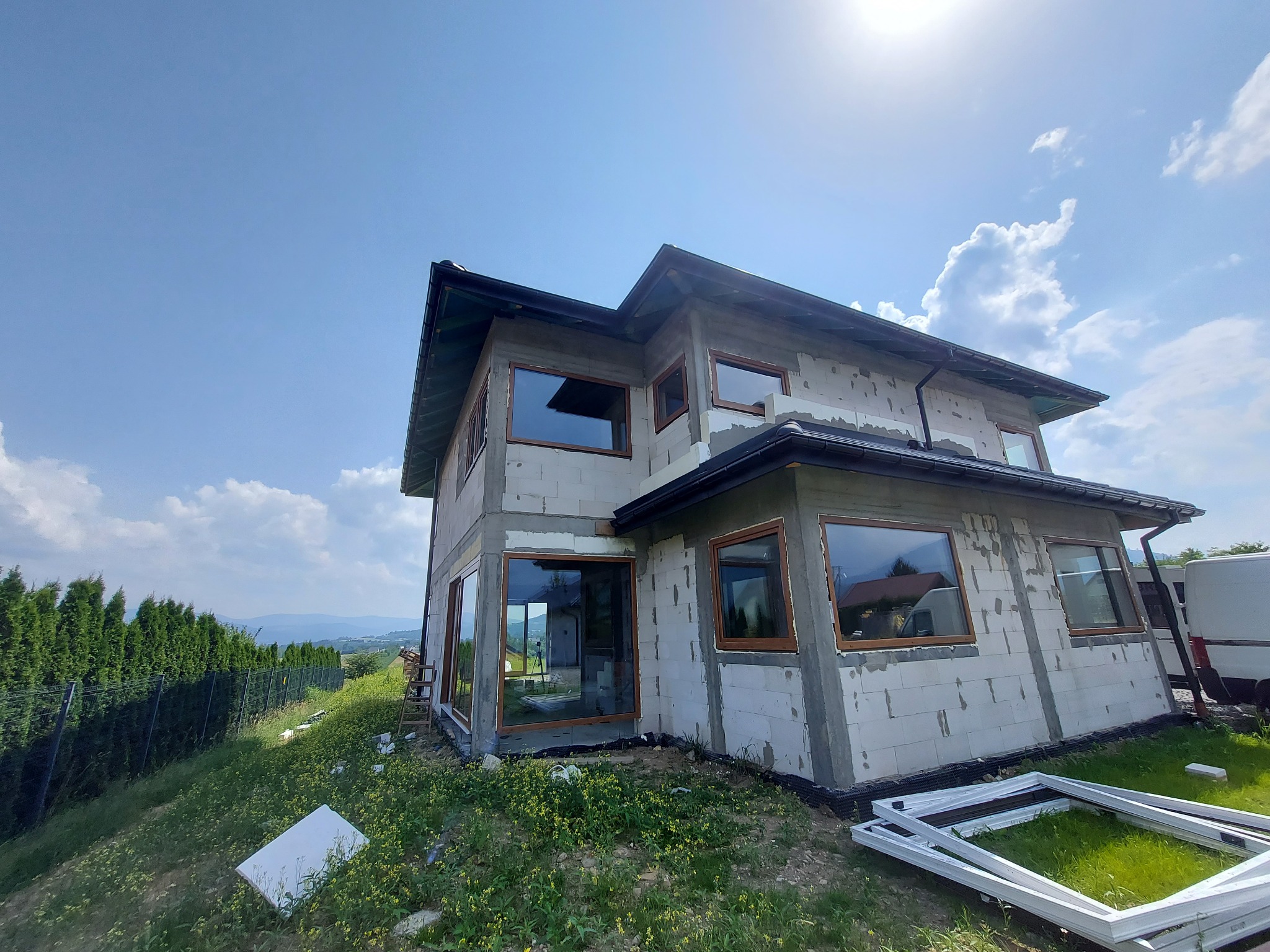 Surowy budynek w trakcie budowy z zamontowanymi oknami o drewnianych ramach, widok z dołu na tle błękitnego nieba z chmurami i zielonego wzgórza.