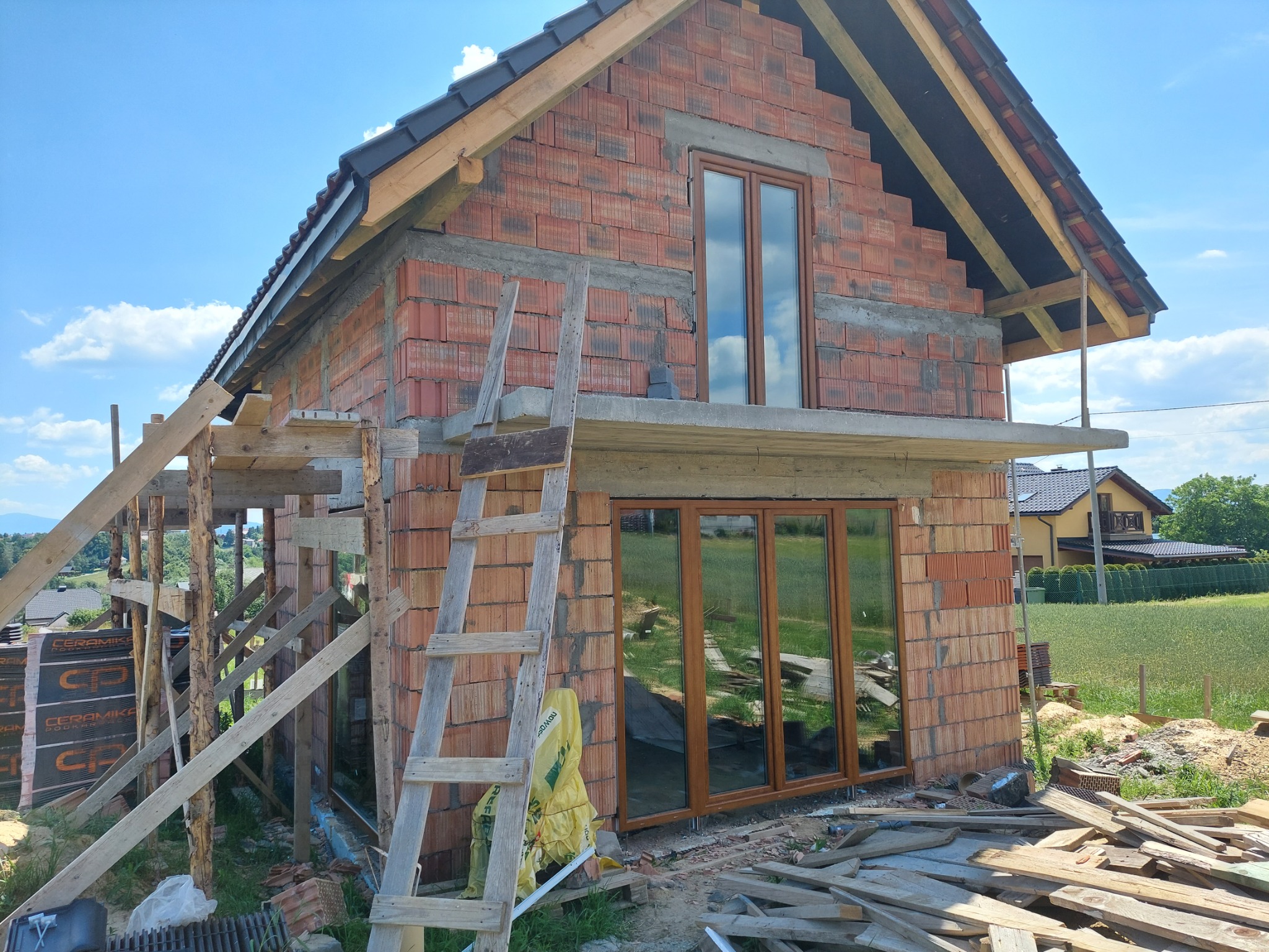 Budowa domu z cegły: wstawione okna na parterze i poddaszu, widoczne rusztowania i drabina, teren budowy z resztkami drewna i materiałów budowlanych.