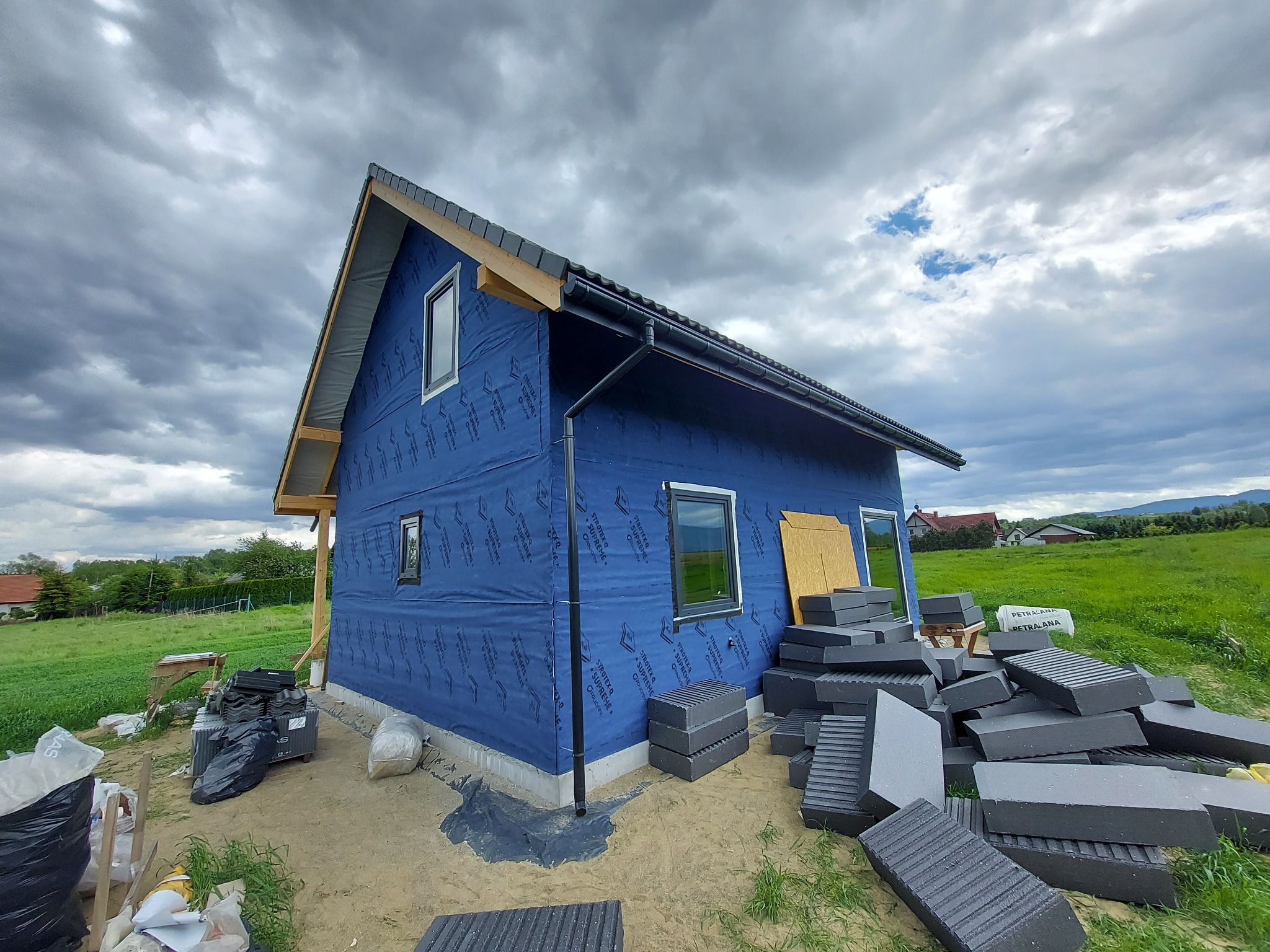 Dom w trakcie budowy w Sopotni Małej, z widoczną niebieską membraną paroprzepuszczalną na ścianach zewnętrznych, nowymi oknami z czarnymi ramami i stertą bloczków izolacyjnych na ziemi.