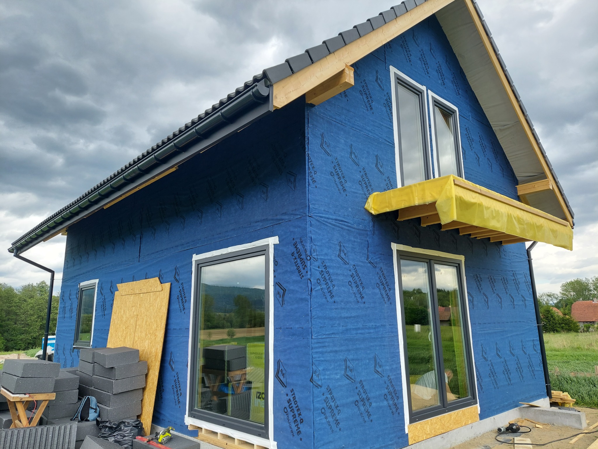 Dom w trakcie ocieplania, widoczne okna z szarymi ramami, niebieska membrana wiatroizolacyjna z nadrukiem, przygotowane bloczki fundamentowe i płyta OSB oparta o ścianę.
