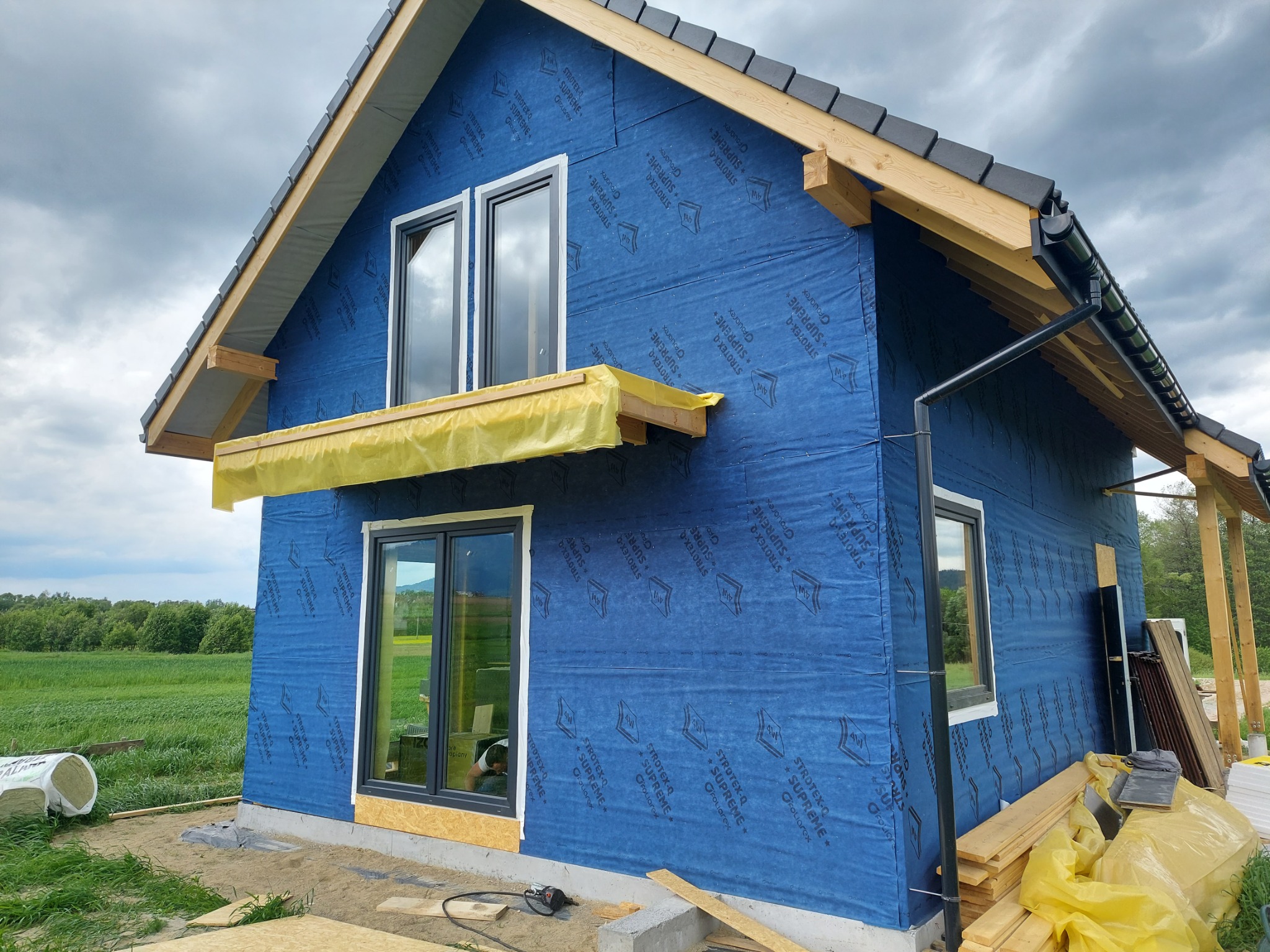 Dom w trakcie budowy z oknami, zabezpieczony niebieską folią paroizolacyjną z nadrukiem, widoczne materiały budowlane i narzędzia na zewnątrz.