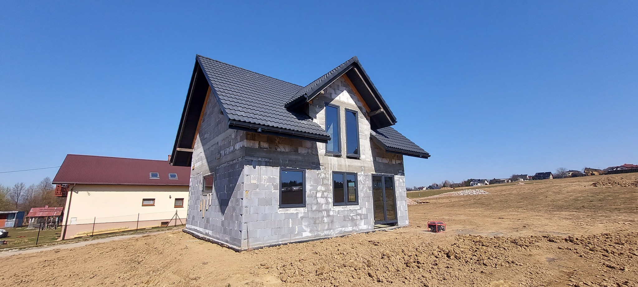 Dom w stanie surowym otwartym z zamontowanymi oknami i dachem z dachówki ceramicznej, widoczny agregat prądotwórczy.