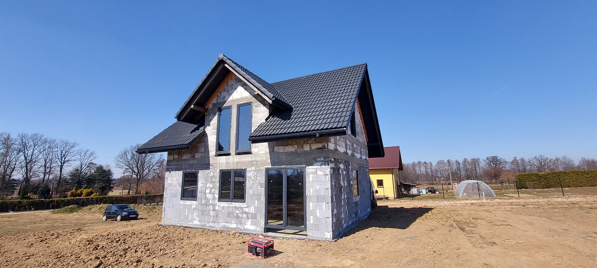 Surowy stan domu jednorodzinnego z szarą dachówką, widoczne okna i drzwi w ciemnych ramach, teren wokół domu w stanie przygotowania pod ogród, w tle drugi budynek i drzewa.