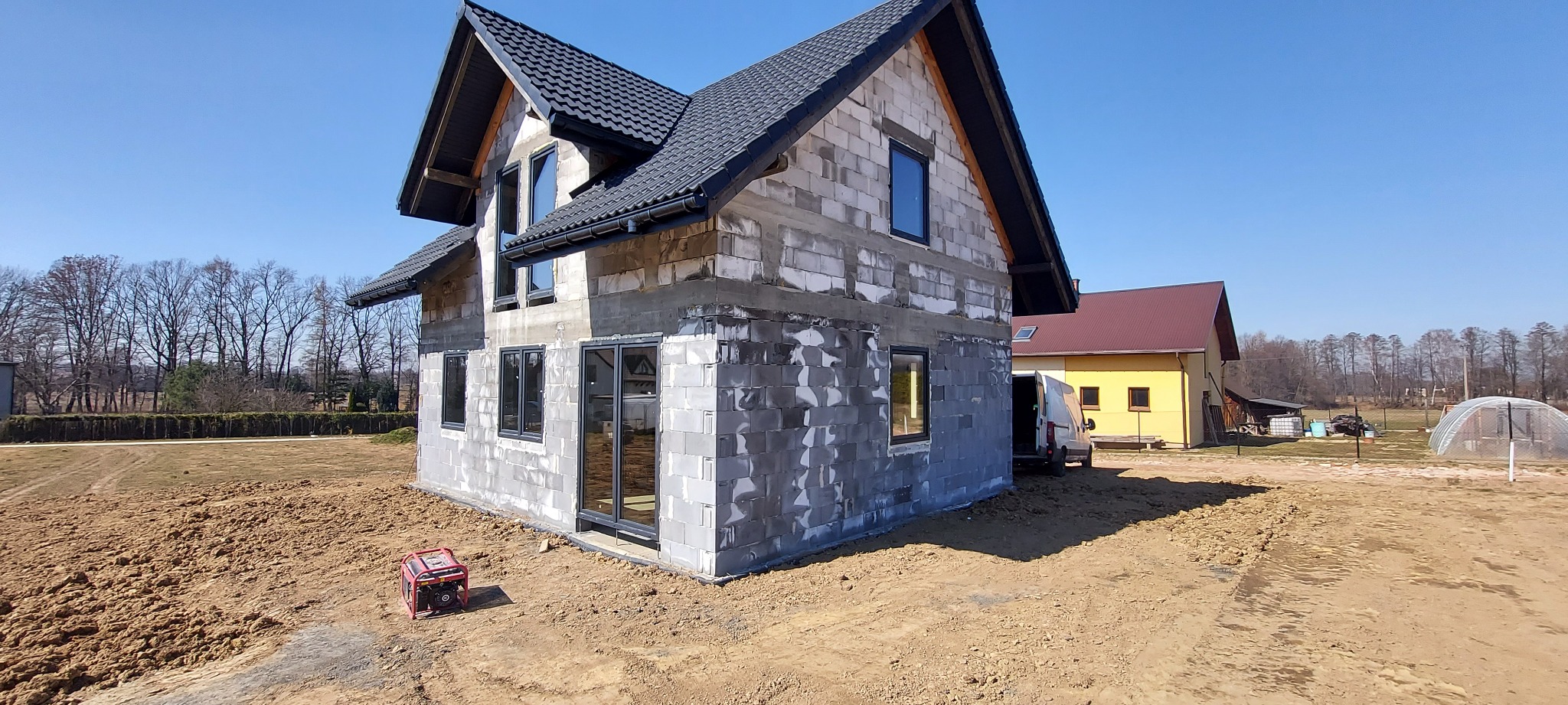 Surowy stan domu jednorodzinnego z szarej cegły, z zamontowanymi czarnymi oknami i drzwiami, na placu budowy z widocznym agregatem prądotwórczym.