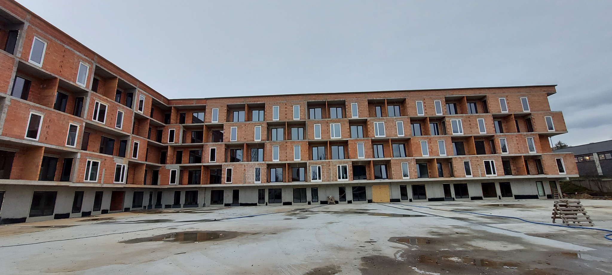 Wielopiętrowy budynek w trakcie budowy, z częściowo zamontowanymi oknami i drzwiami, widoczne surowe mury z czerwonej cegły i betonowa podstawa, plac budowy z kałużami.