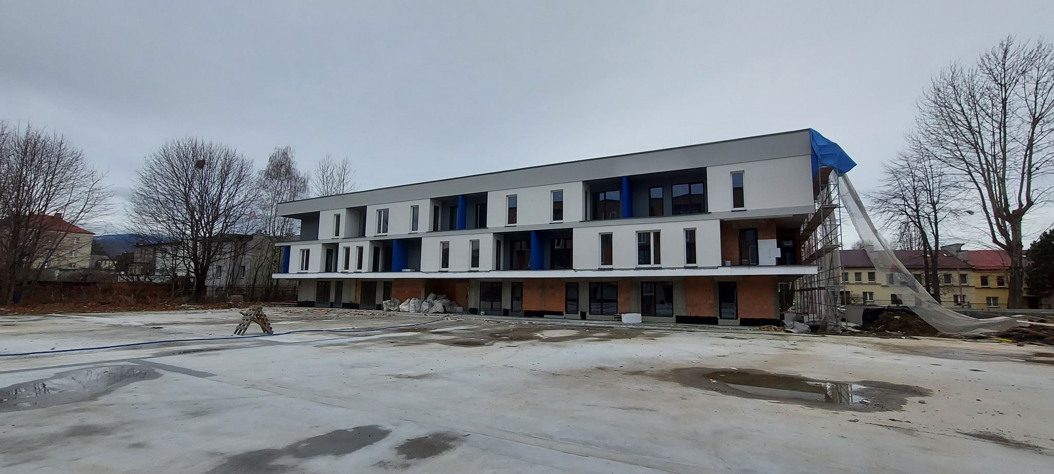 Wielorodzinny budynek w trakcie budowy, widoczne rusztowania, niezakończone elewacje, okna zamontowane w części budynku, plac budowy z workami i materiałami budowlanymi.