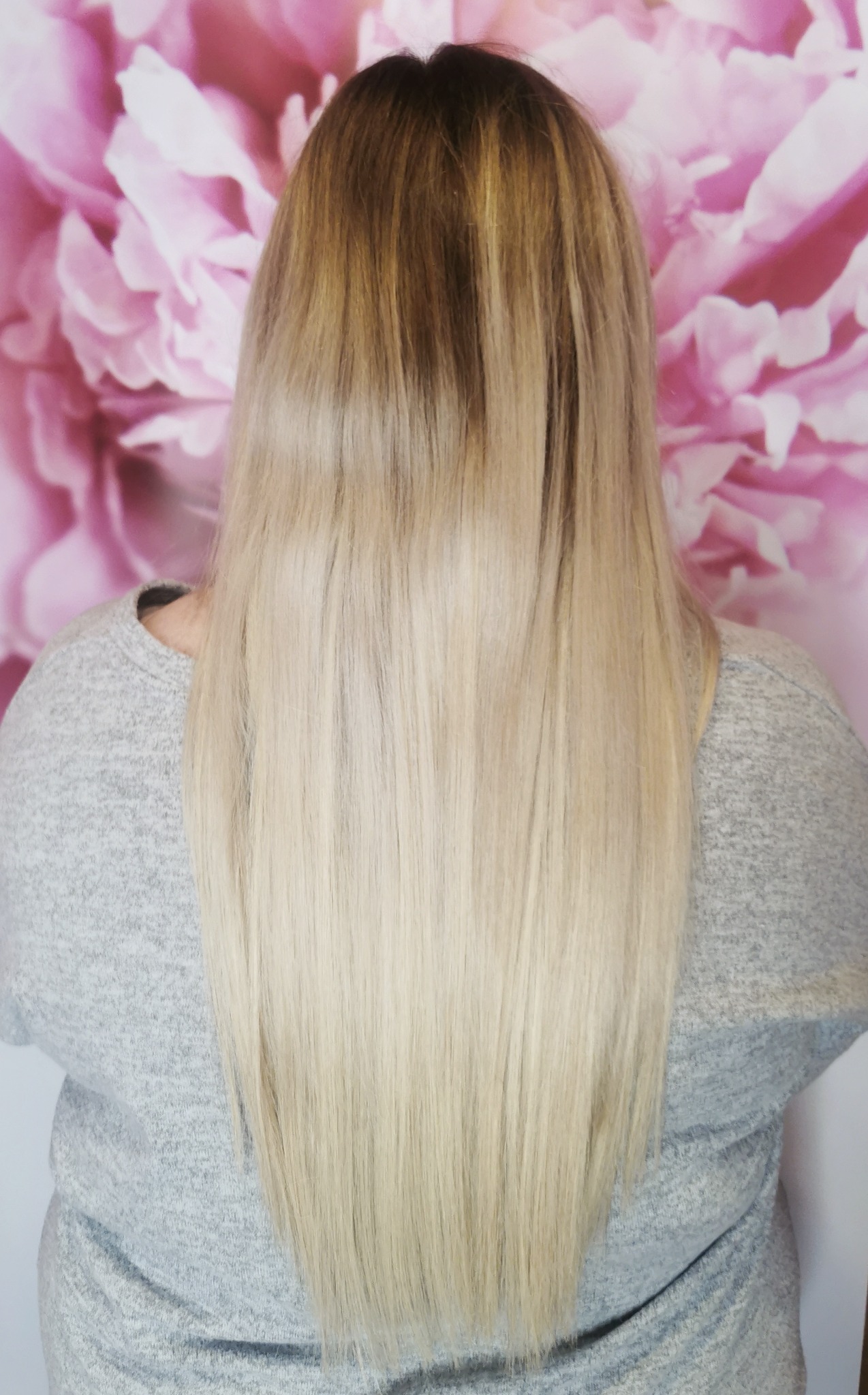 Długie, proste włosy z ombre, przechodzące od ciemnego blondu u nasady do jasnego blondu na końcach, na tle ściany z motywem różowych piwonii.