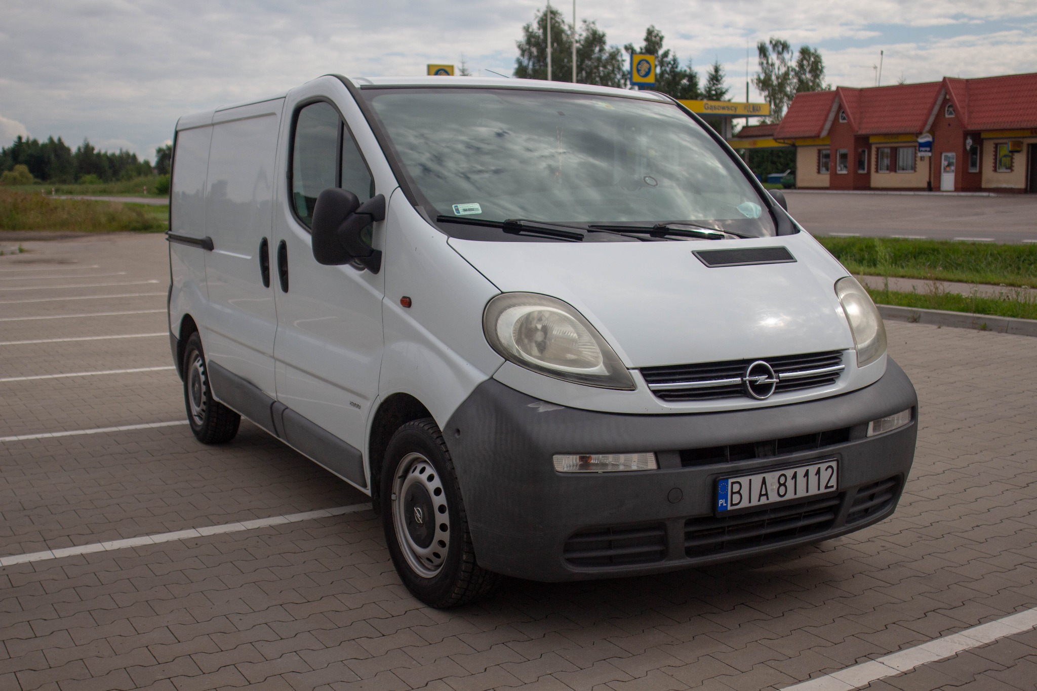 Biały Opel Vivaro zaparkowany na parkingu z kostki brukowej, widoczny budynek stacji benzynowej w tle.
