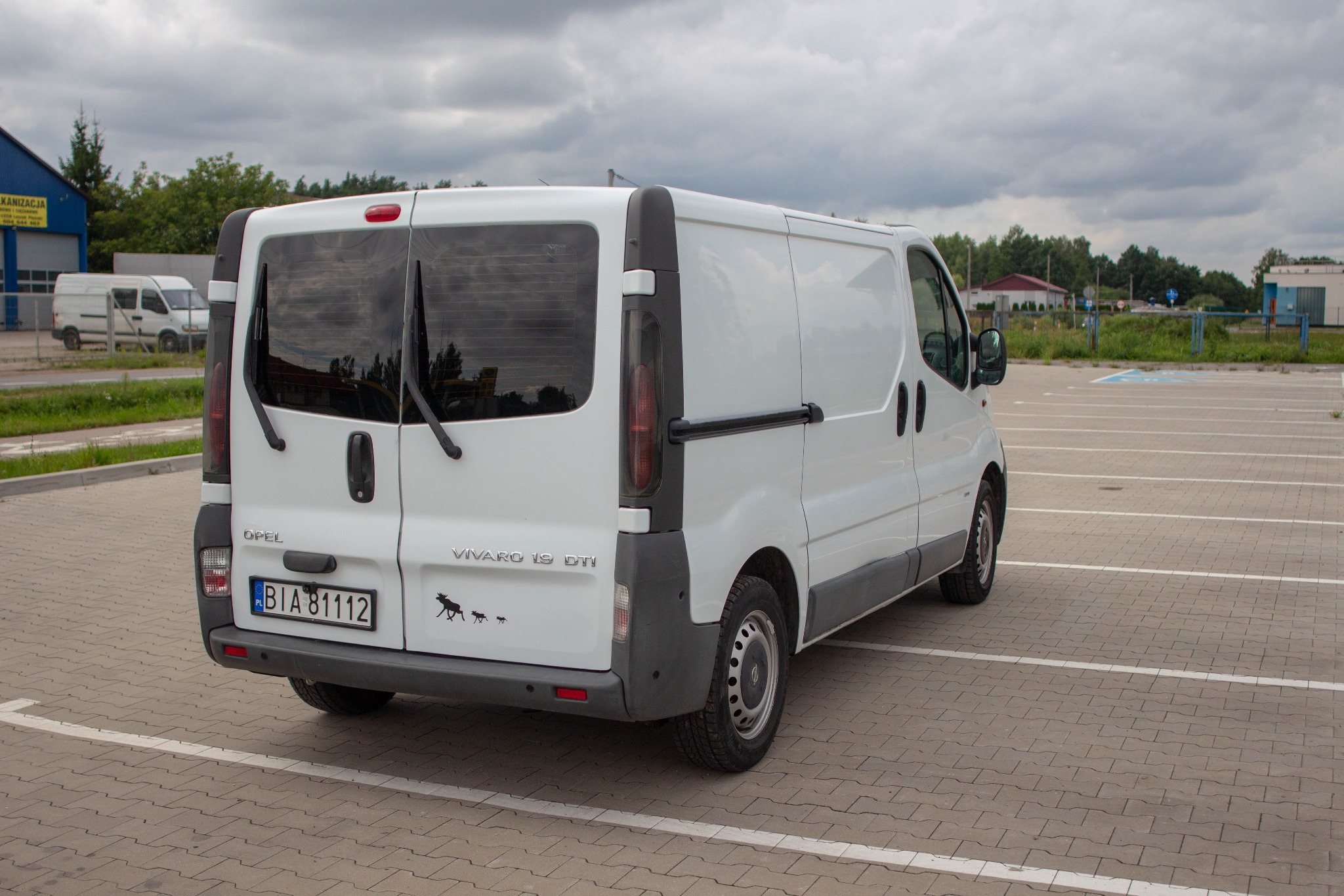 Biały Opel Vivaro 1.9 DTI zaparkowany na parkingu z kostki brukowej, widok z tyłu, na zderzaku naklejki z motywem łosia i wilków.