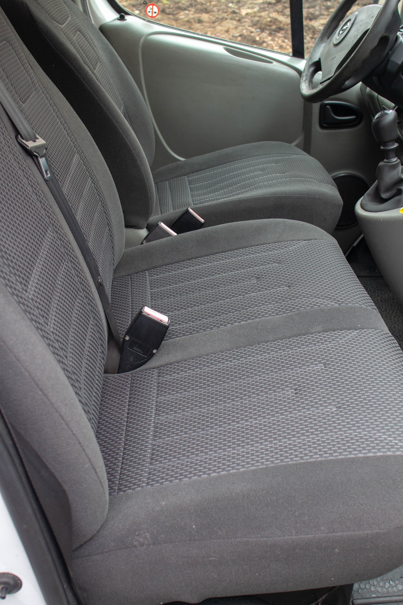 Wnętrze samochodu dostawczego: szare fotele z pasami bezpieczeństwa, widoczna kierownica i dźwignia zmiany biegów.