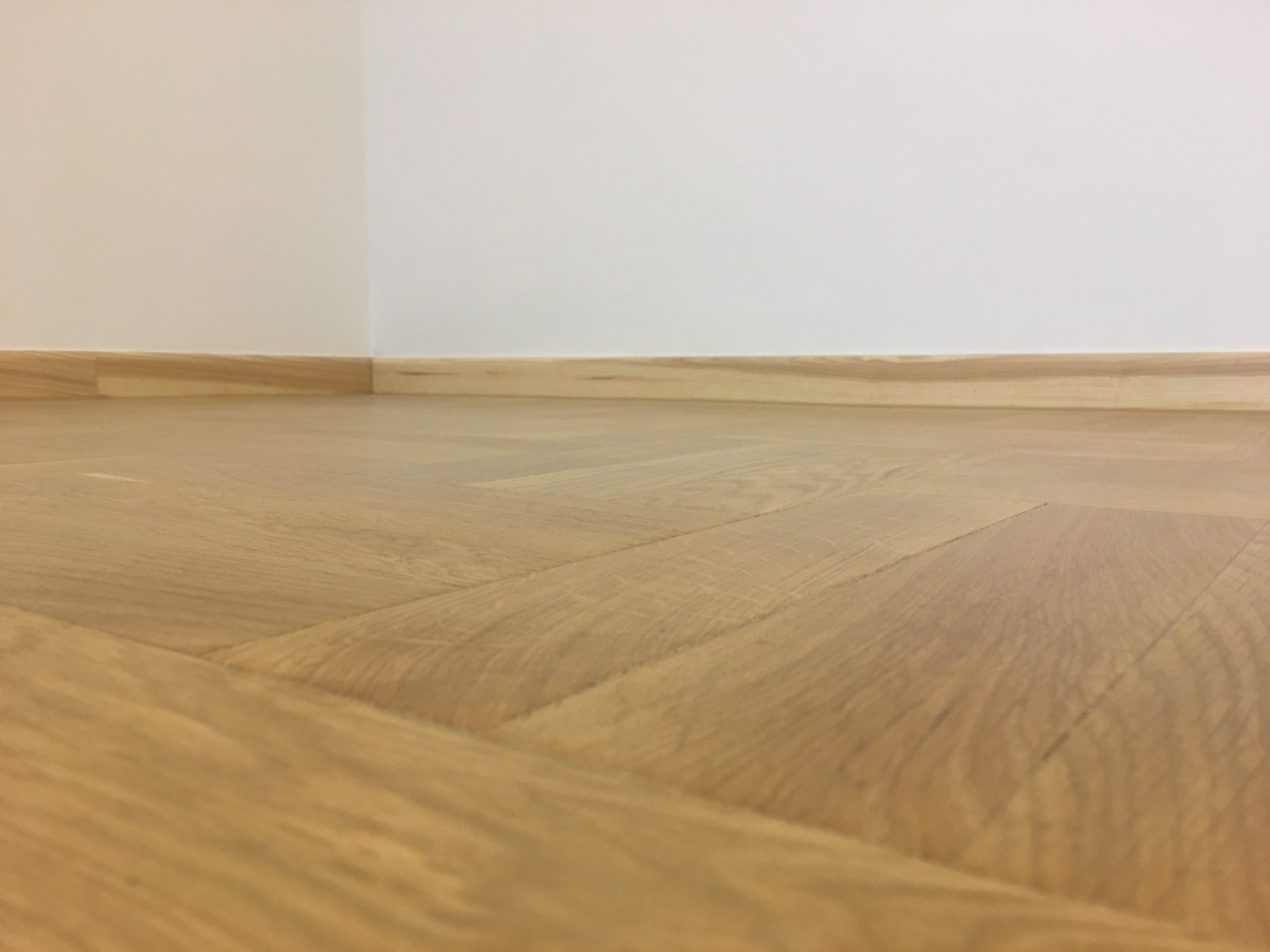 Parkiet ułożony w jodełkę, przy krawędzi ściany z zamontowaną drewnianą listwą przypodłogową.