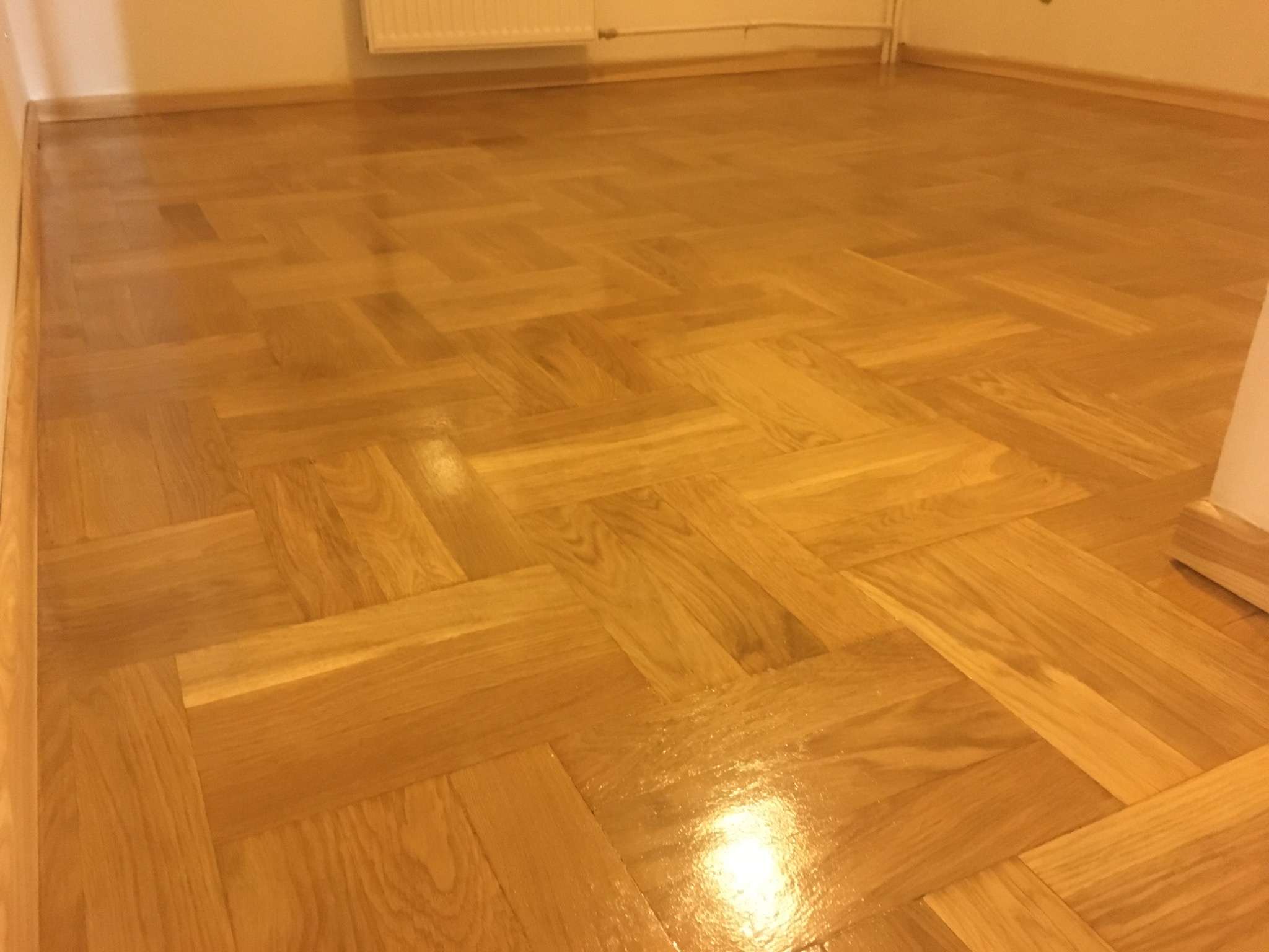 Parkiet ułożony w jodełkę po cyklinowaniu i lakierowaniu, widoczny połysk na powierzchni, listwy przypodłogowe w kolorze drewna.