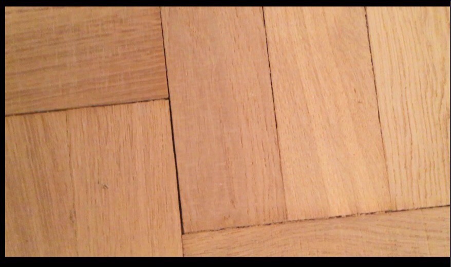 Parkiet ułożony w jodełkę, widoczne rysy i drobne zabrudzenia w szczelinach.