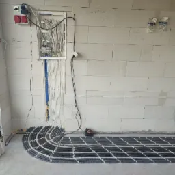 Rozprowadzenie kabli elektrycznych w budynku w stanie surowym, widoczna skrzynka rozdzielcza z okablowaniem, przewody ułożone w rzędach na podłodze, ściany z betonu komórkowego.