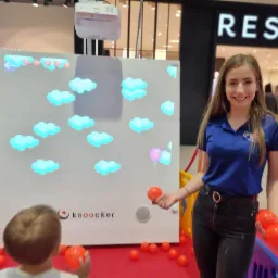 Uśmiechnięta animatorka w niebieskiej koszulce prezentuje interaktywną tablicę z projekcją chmur i balonów, otoczoną kolorowymi piłkami, podczas eventu w centrum handlowym.