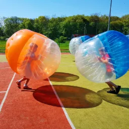 Dzieci w przezroczystych kulach, jedna pomarańczowa, druga niebieska, na boisku z zielono-czerwonym podziałem, w tle drzewa i niebieskie niebo.