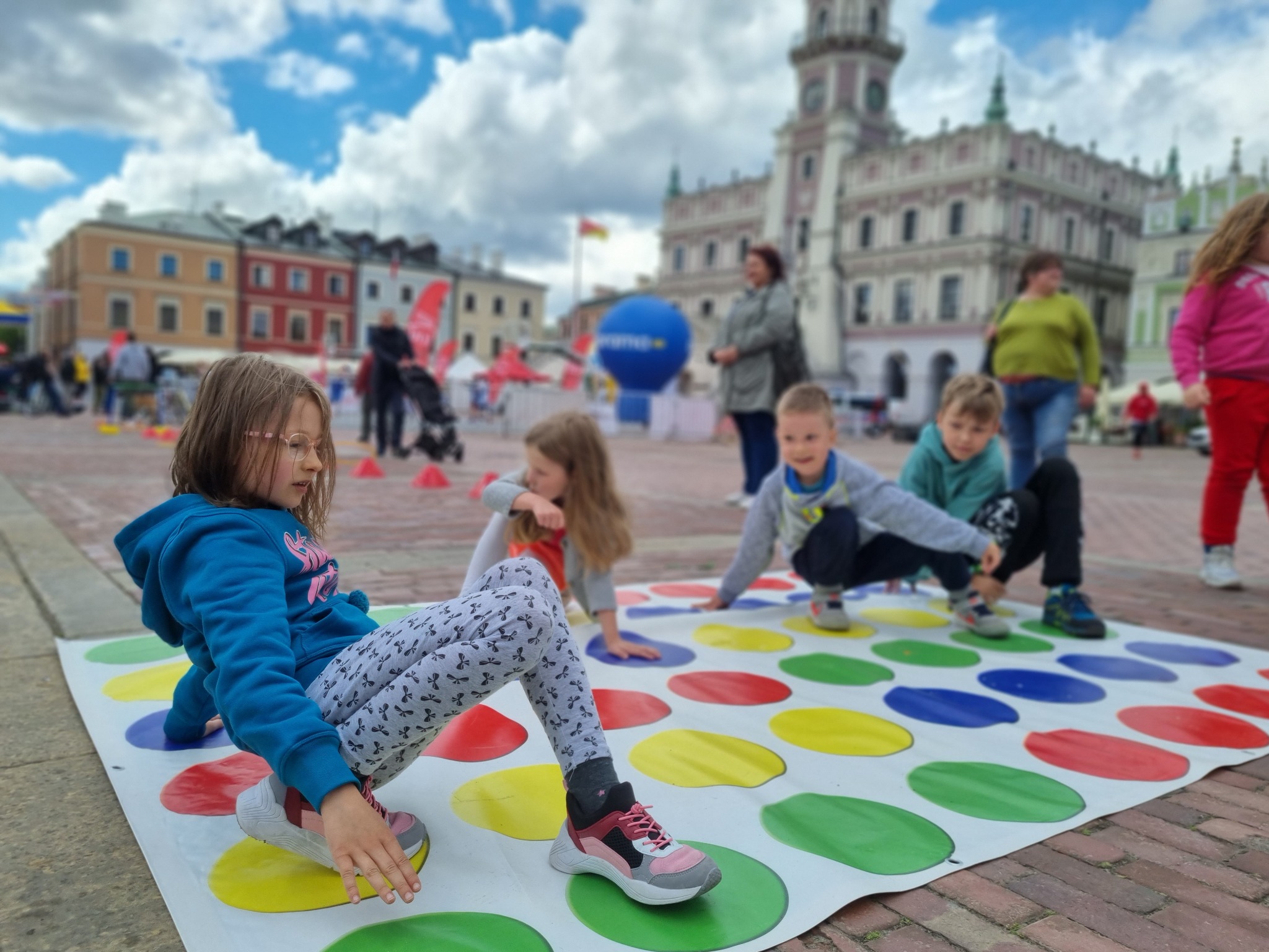 Dzieci grające w Twistera na placu miejskim z zabytkowym ratuszem w tle, widoczne kolorowe kropki i pozy graczy.