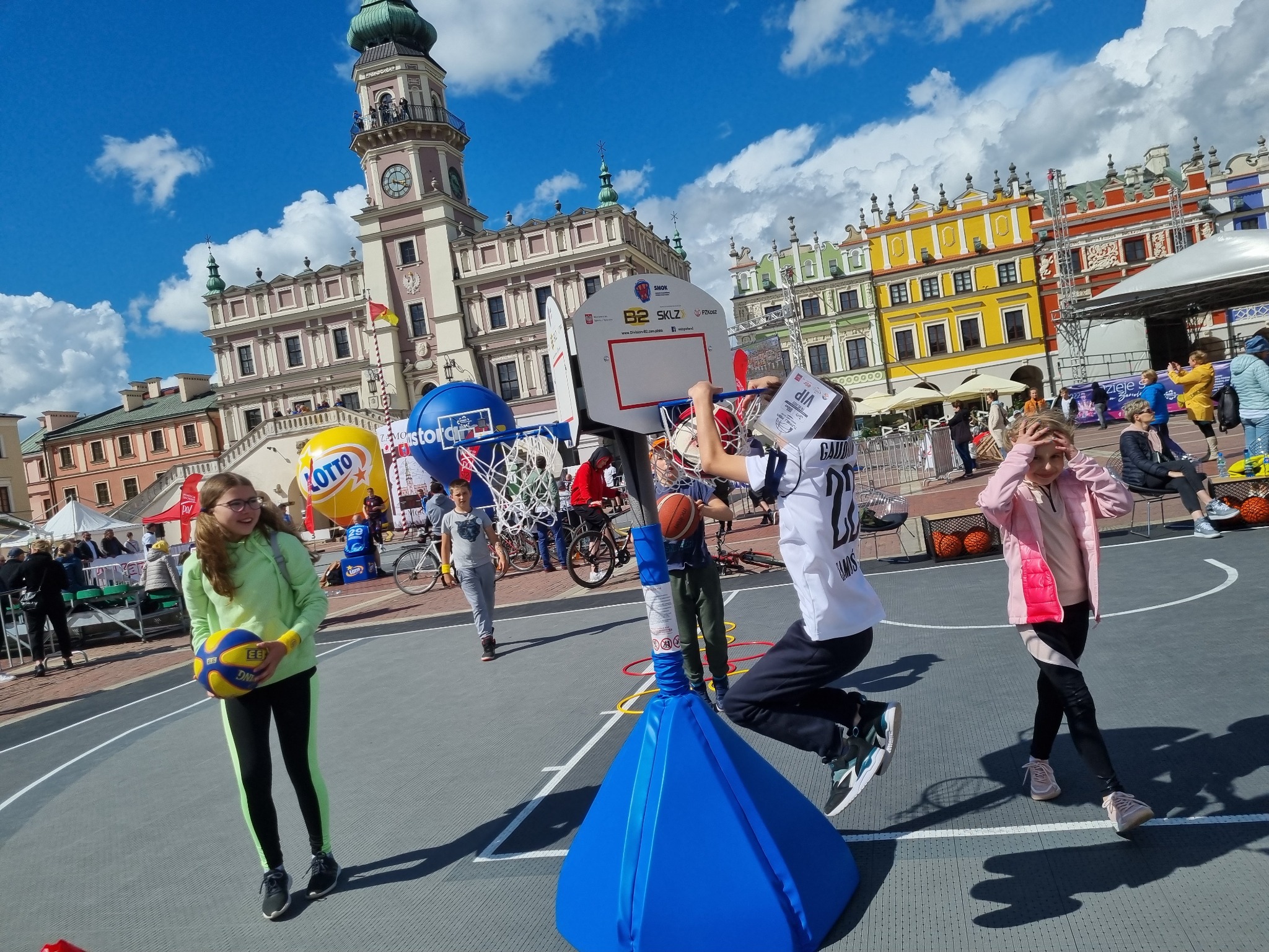 Dzieci uczestniczą w sportowej imprezie plenerowej na rynku miasta Zamość, z dmuchanymi balonami Lotto w tle, podczas gdy chłopiec w koszulce sportowej skacze do kosza.