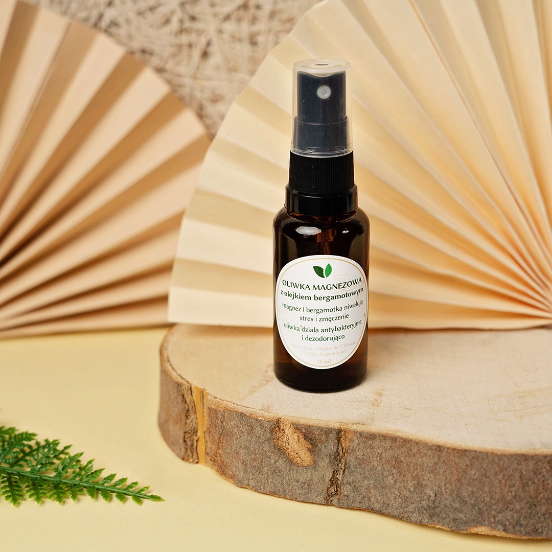 Butelka oliwki magnezowej z olejkiem bergamotowym na drewnianym krążku, z wachlarzami w tle i zieloną gałązką w lewym dolnym rogu.