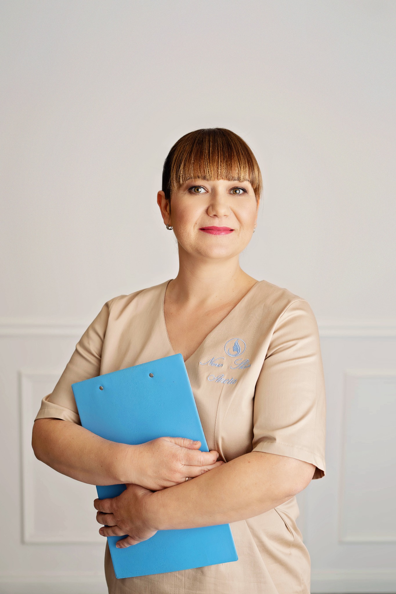 Portret kobiety w beżowym uniformie z logo, trzymającej niebieski clipboard na tle białej ściany.