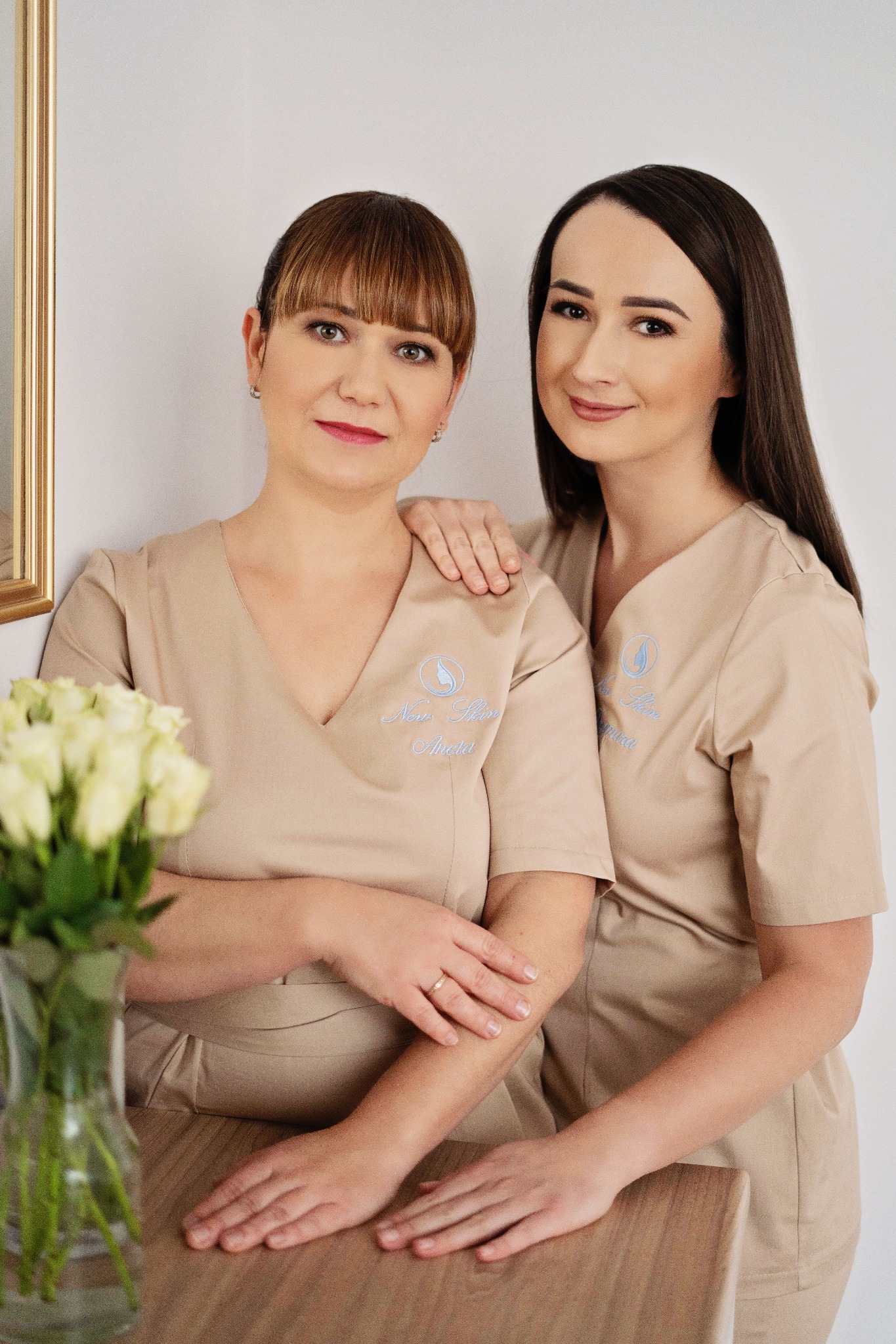 Dwie kobiety w beżowych uniformach z logo firmy, pozujące na tle białej ściany z bukietem białych róż w wazonie, uśmiechnięte i patrzące w obiektyw.