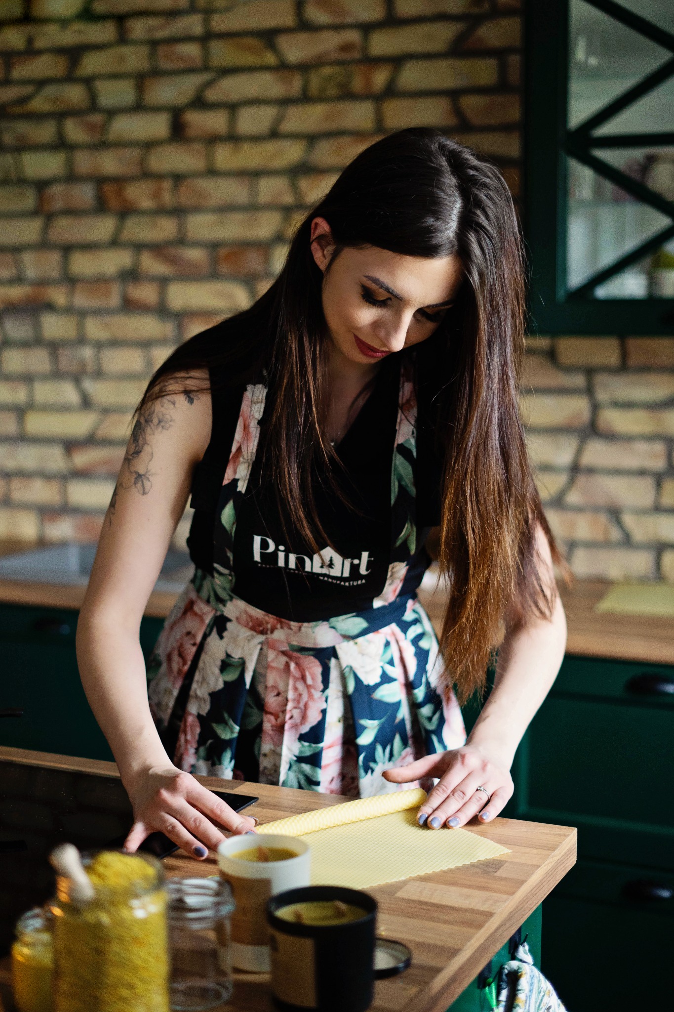 Kobieta w kwiecistej spódnicy i fartuchu z logo, pochylona nad drewnianym blatem, wyrównuje arkusz wosku pszczelego, obok słoiki i kubki z woskiem, ceglana ściana w tle.