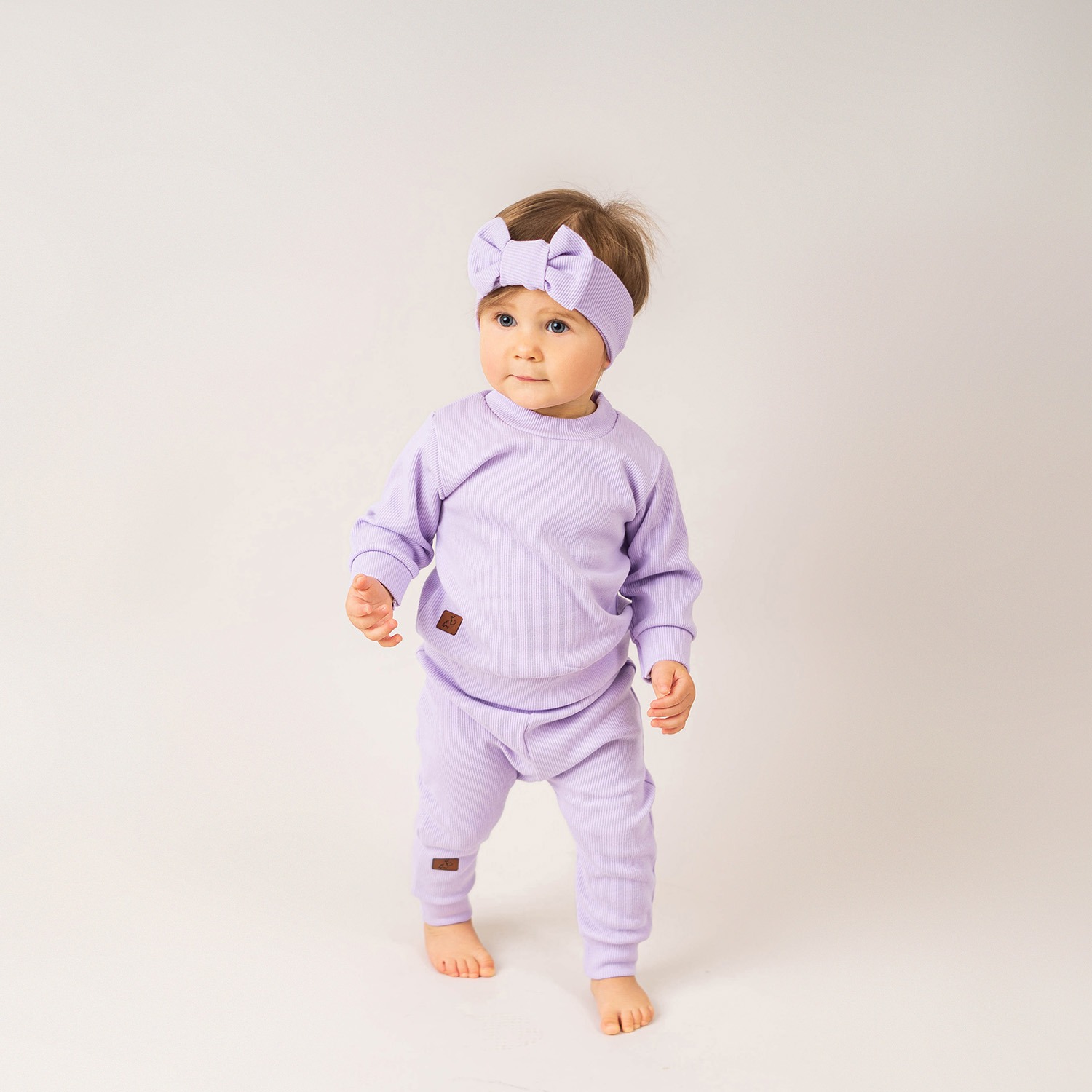 Urocza dziewczynka w prążkowanym liliowym komplecie z kokardą na głowie, stojąca na białym tle i spoglądająca w lewo.