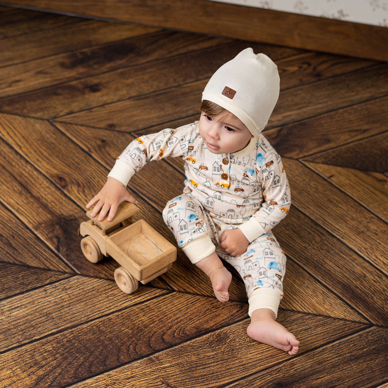 Mały chłopiec w piżamie z motywem domków i autek siedzi na drewnianej podłodze i bawi się drewnianą zabawką ciężarówką.