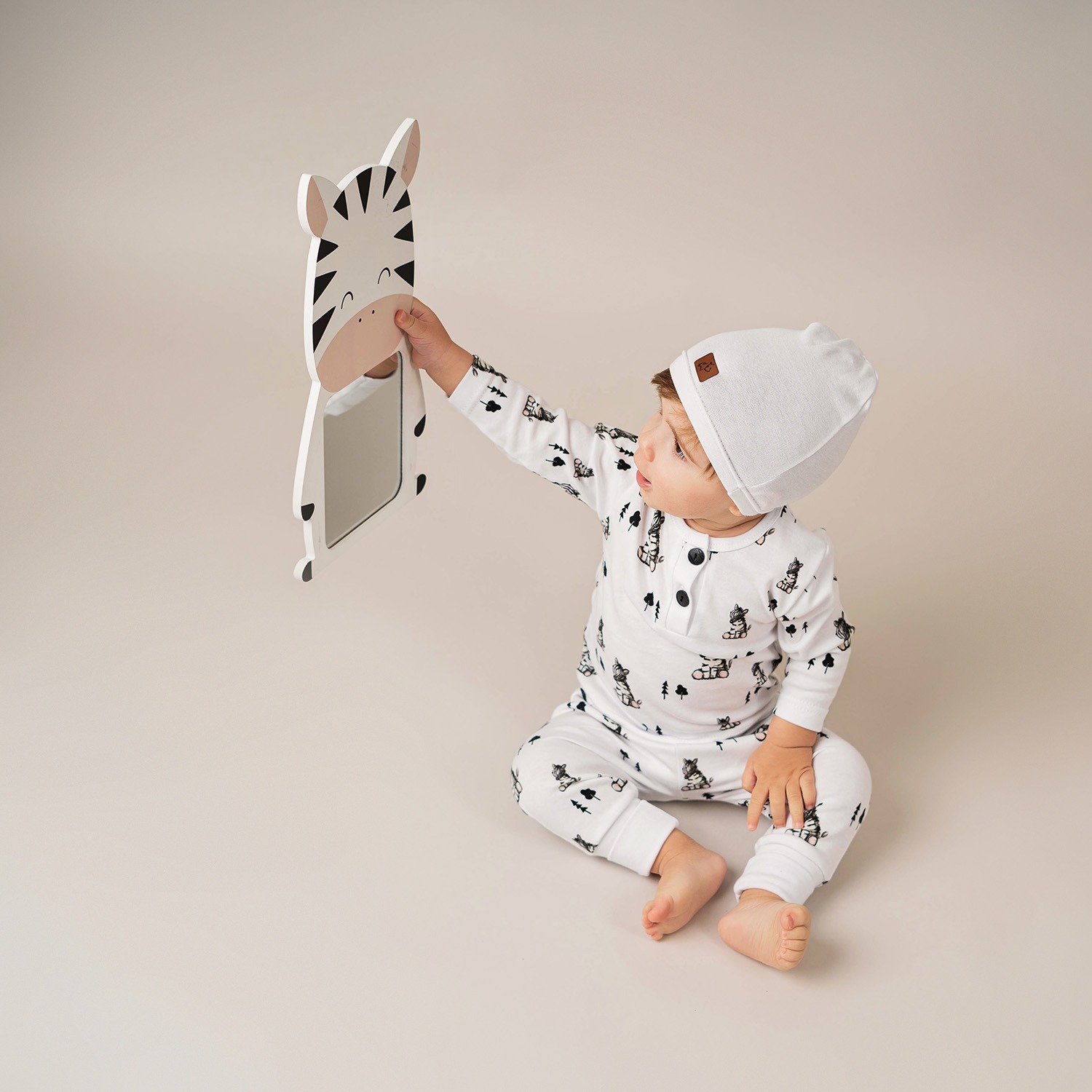 Mały chłopiec w piżamie z motywem zwierzęcym siedzi na podłodze i trzyma lustro w kształcie zebry.