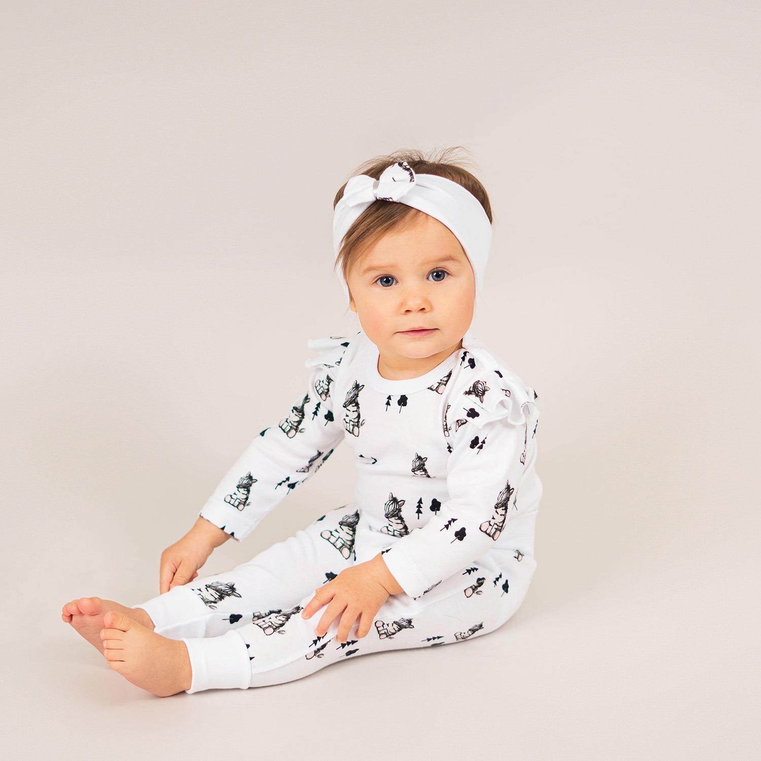 Urocza dziewczynka w białym komplecie z długim rękawem i spodniami, ozdobionym wzorem zwierząt i drzew, siedzi na jasnym tle, ubrana w dopasowaną opaskę z kokardką.