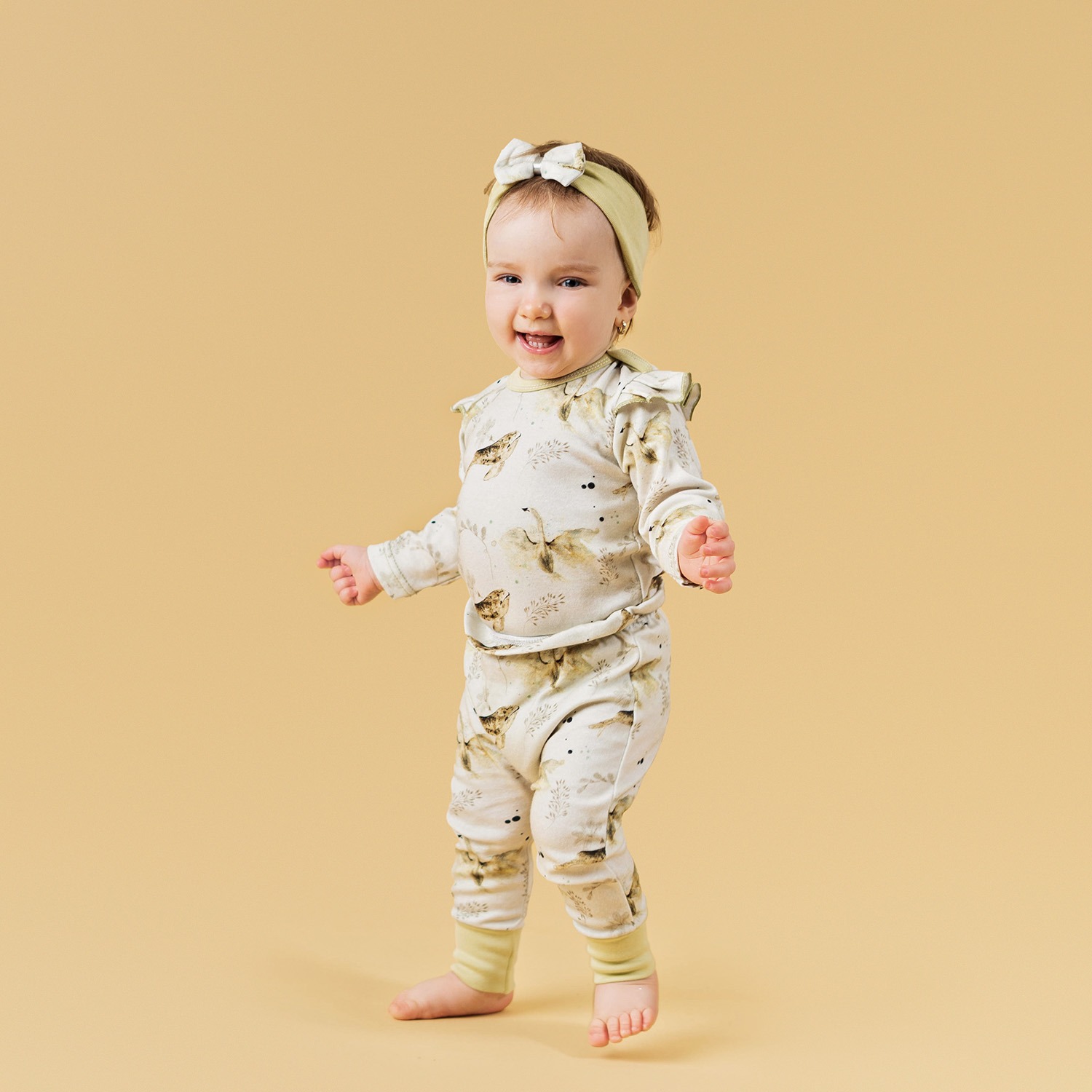 Uśmiechnięta roczna dziewczynka w piżamie w zwierzęta, z opaską na głowie, stawia pierwsze kroki na beżowym tle.