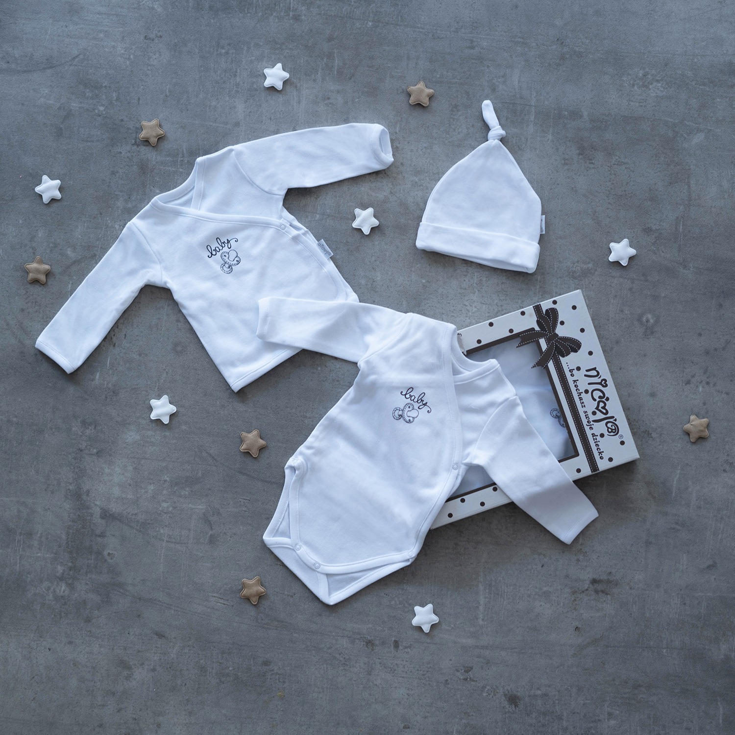 Dwa białe body niemowlęce z haftem, czapeczka z supełkiem i pudełko prezentowe na szarym tle z dekoracyjnymi gwiazdkami.