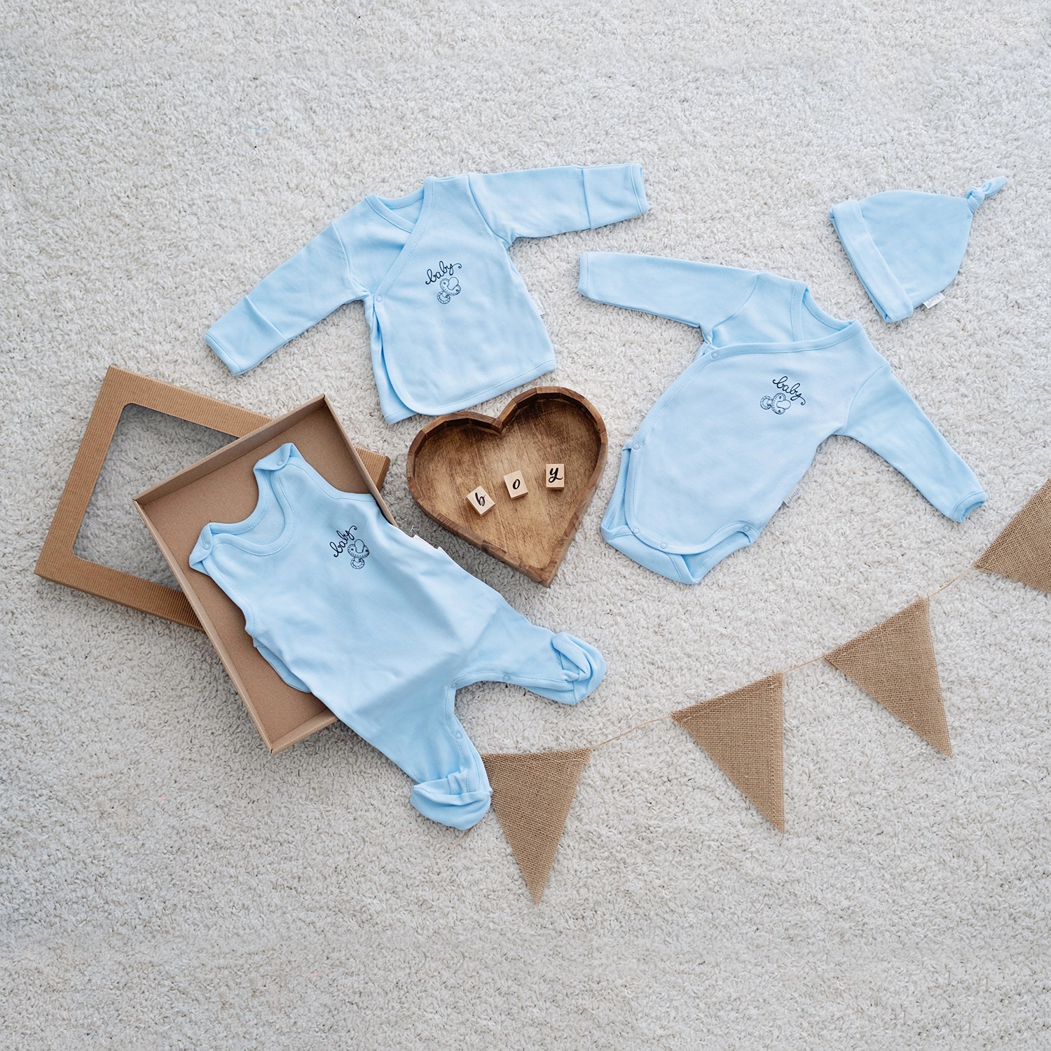 Niebieski zestaw ubranek niemowlęcych z napisem 'baby' i rysunkiem misia, ułożony na puszystym, kremowym dywanie obok drewnianej miseczki w kształcie serca z literami 'boy', otwartego pudełka...