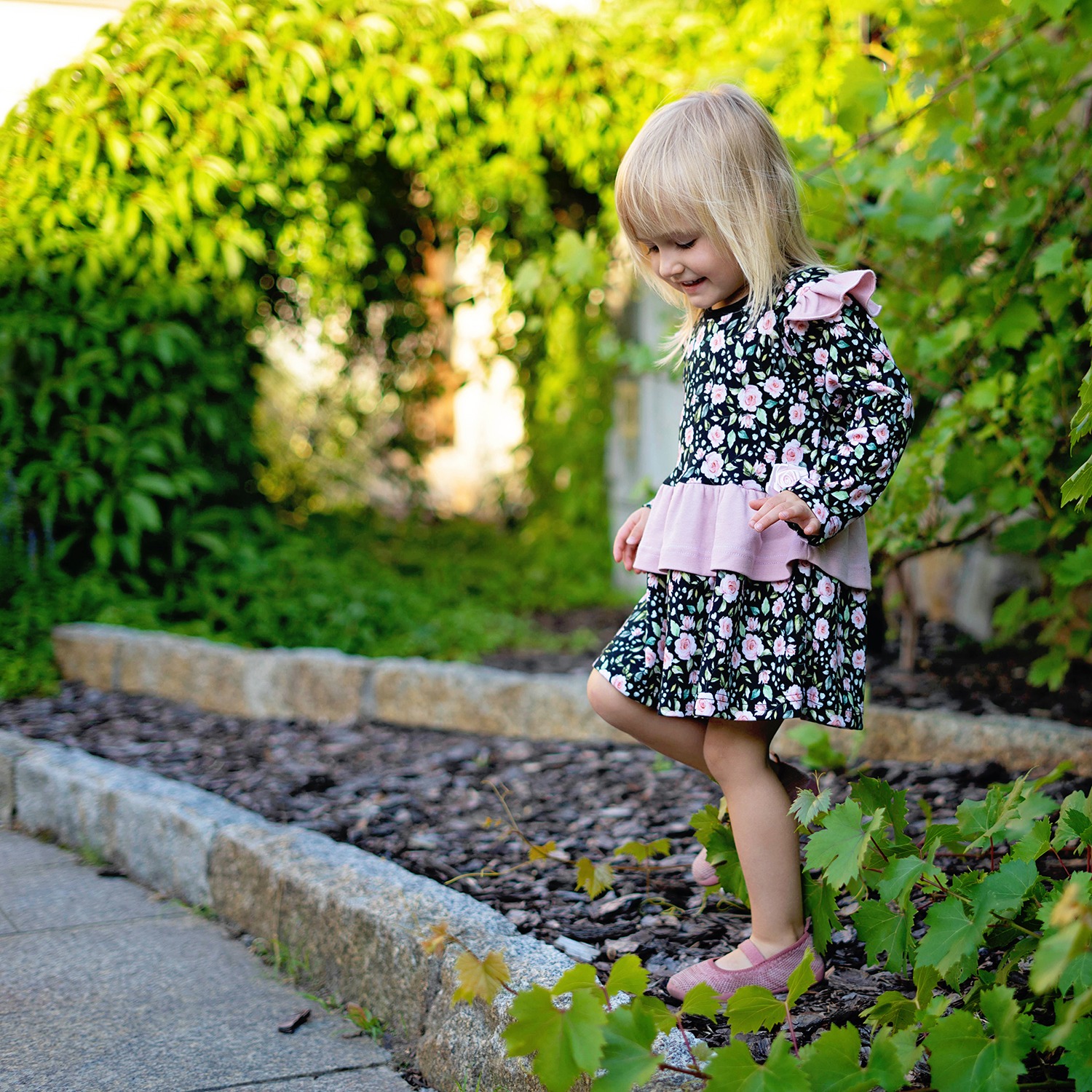 Dziewczynka w kwiecistej sukience i różowych baletkach balansuje na krawężniku w ogrodzie, spoglądając w dół.