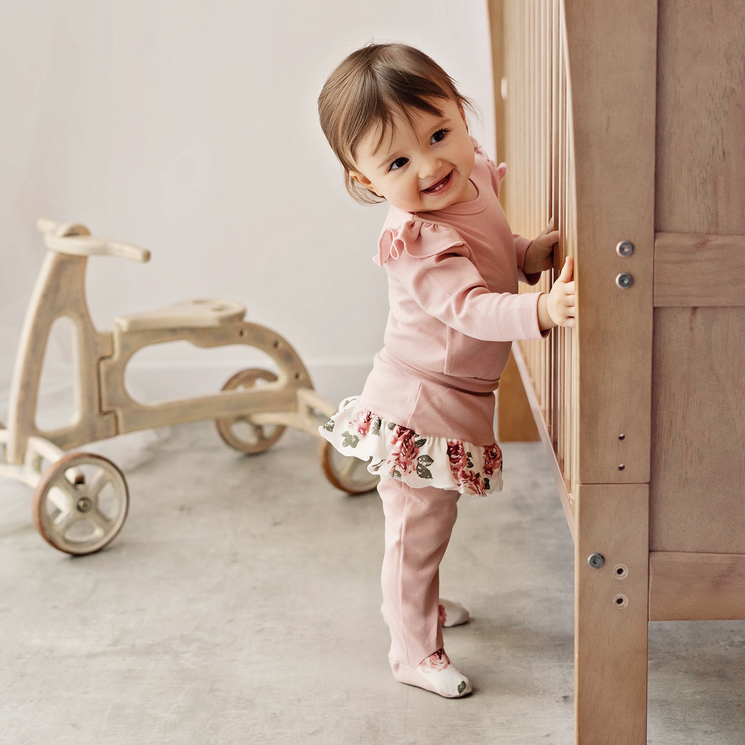 Uśmiechnięta dziewczynka w różowym stroju z falbanką w kwiaty, opierająca się o drewniane łóżeczko dziecięce w Krosno, obok drewniany jeździk.