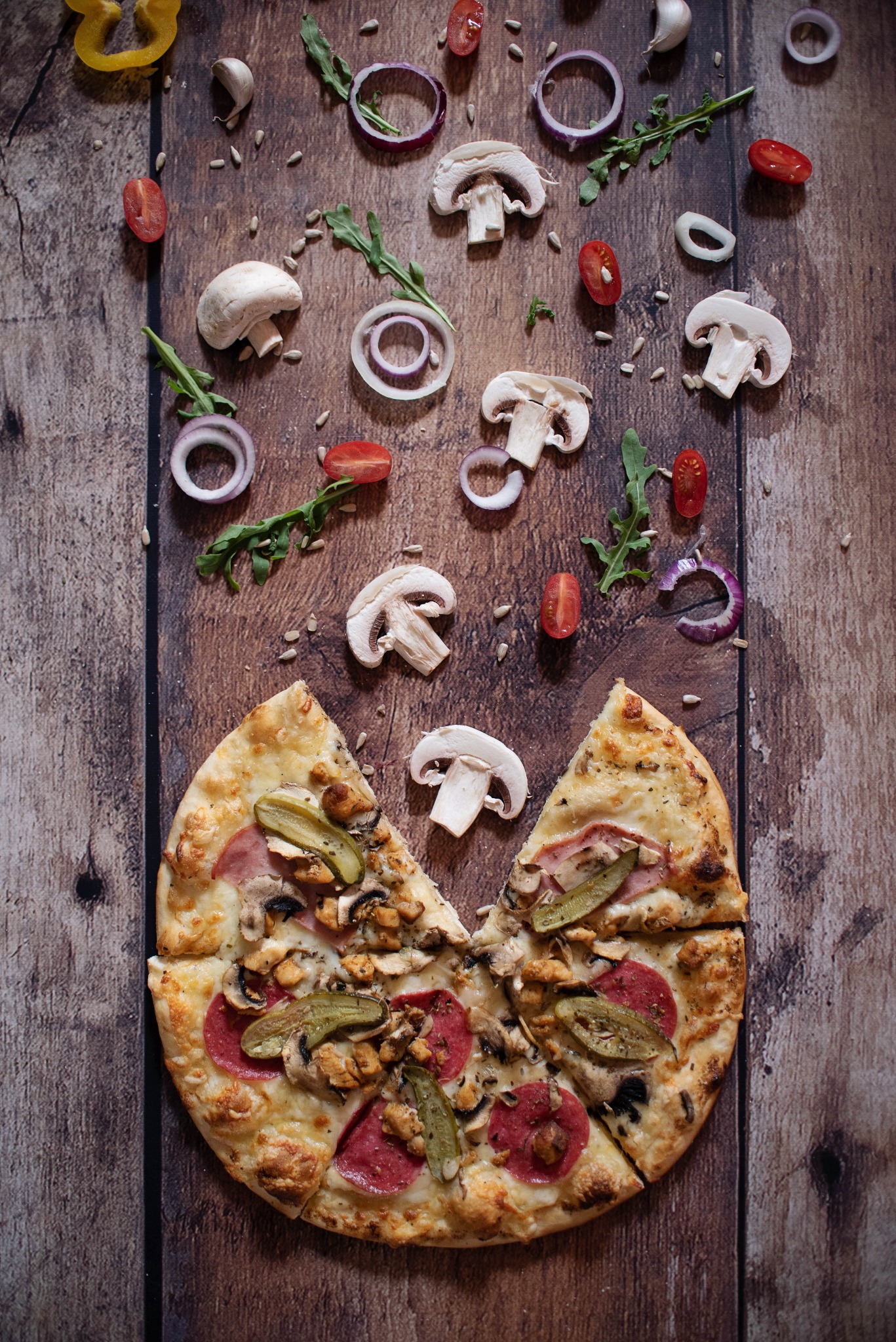 Apetyczna pizza z salami, pieczarkami i ogórkiem konserwowym na drewnianym tle, otoczona luźno rozrzuconymi składnikami: pieczarki, cebula, pomidorki cherry, rukola i ziarna słonecznika.